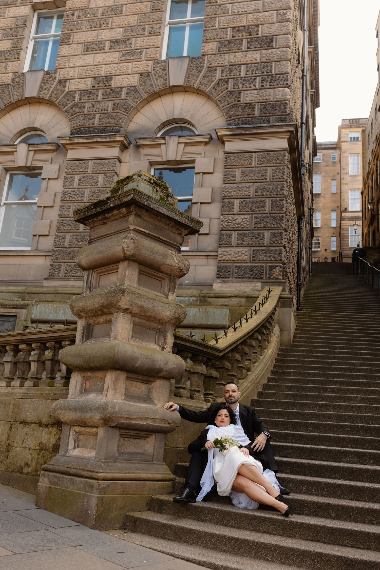 Bride and groom sit relaxed on stone steps beside ornate pillar of a historic sandstone building.