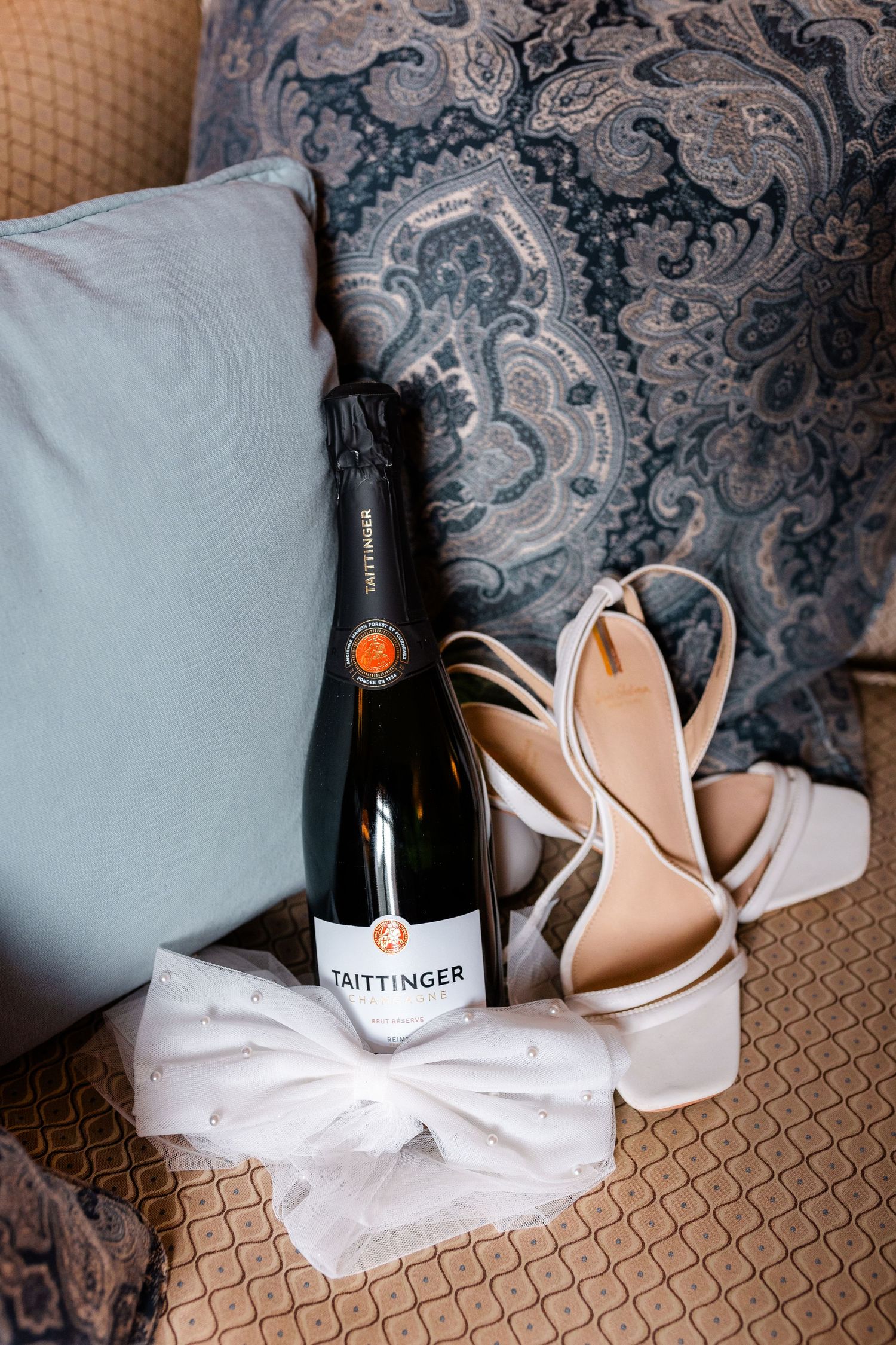Taittinger champagne bottle with white bridal heels and bow on a decorative sofa cushion.