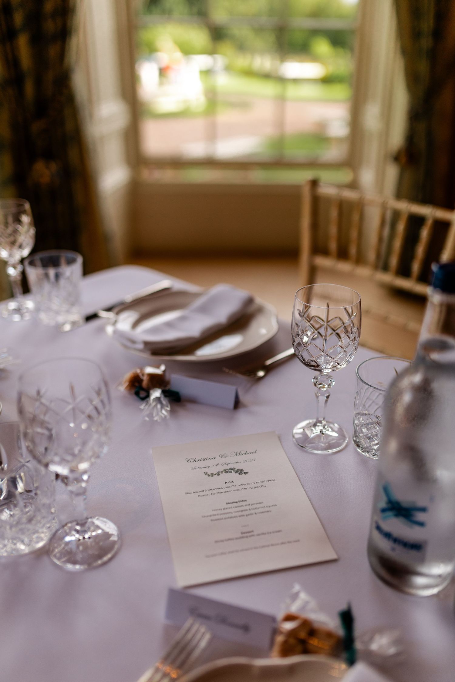 Elegant wedding reception table setting with crystal glassware, white linens, menu cards, and chiavari chairs by window.