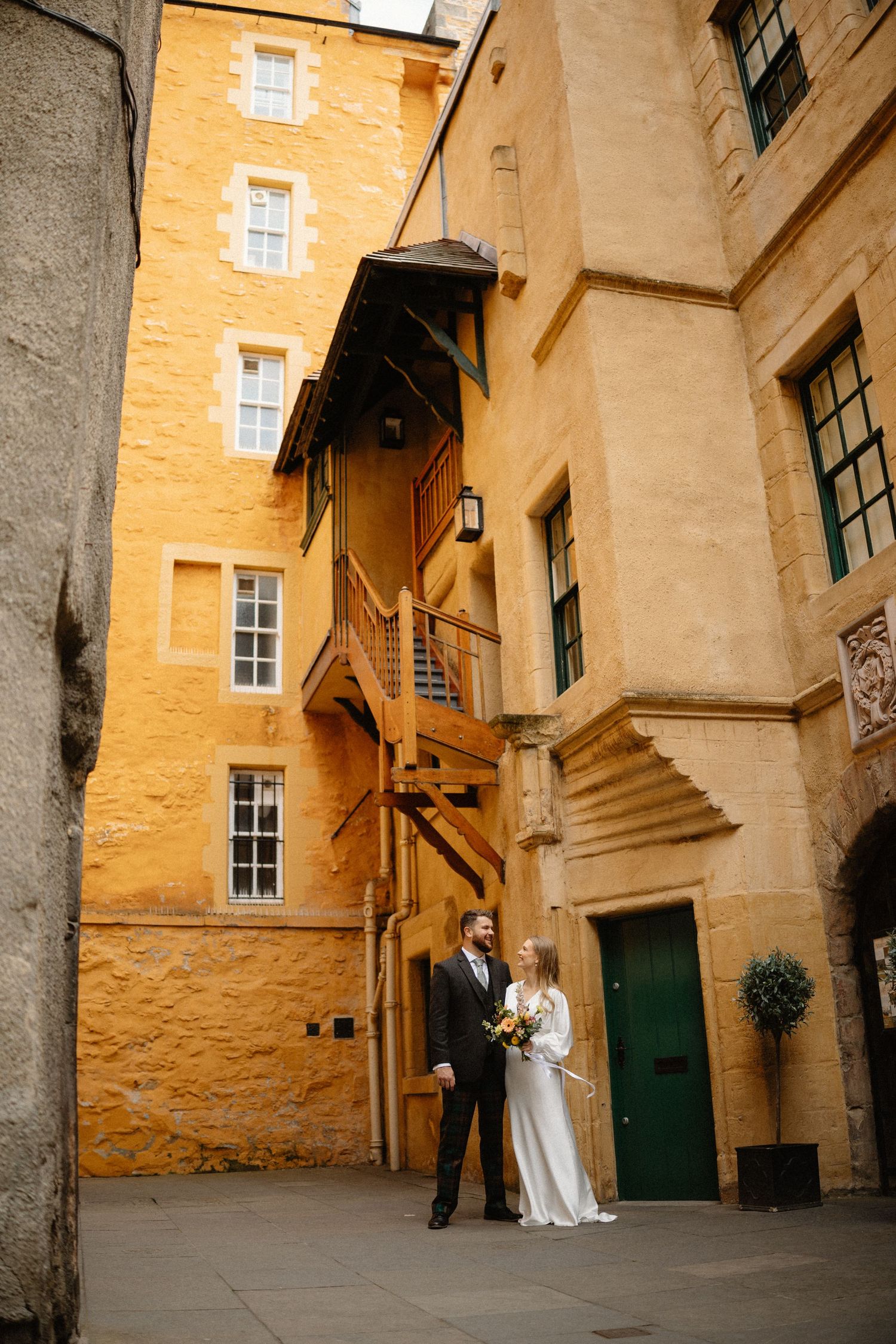 Bride and groom stand in a cobblestone courtyard beside a golden stone building with wooden staircase.