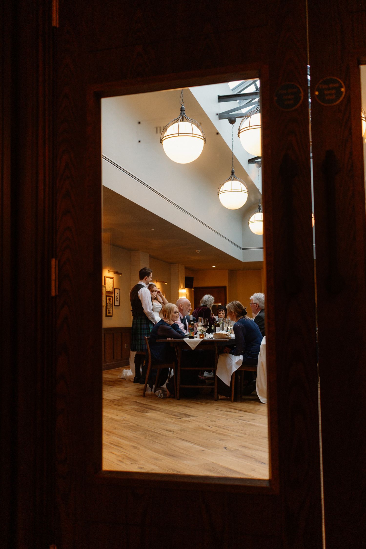 View through wooden door of diners at candlelit restaurant table with globe pendant lights.