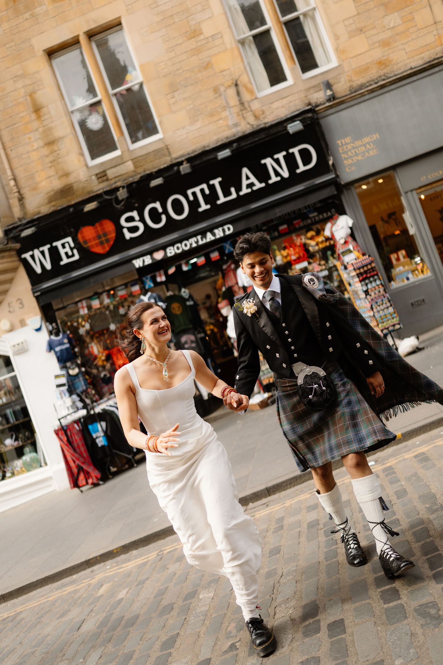 Newlyweds hold hands walking past We Love Scotland shop, bride in white dress, groom in traditional Scottish kilt outfit.