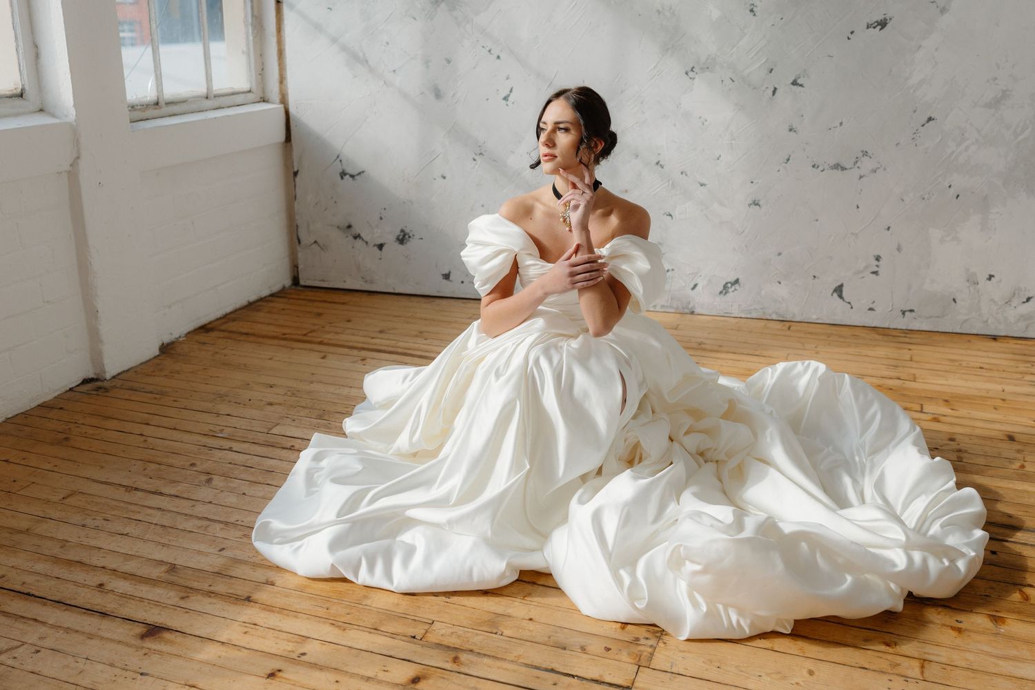 Bride in elegant white ball gown with off-shoulder sleeves sitting gracefully on wooden floor in bright studio.