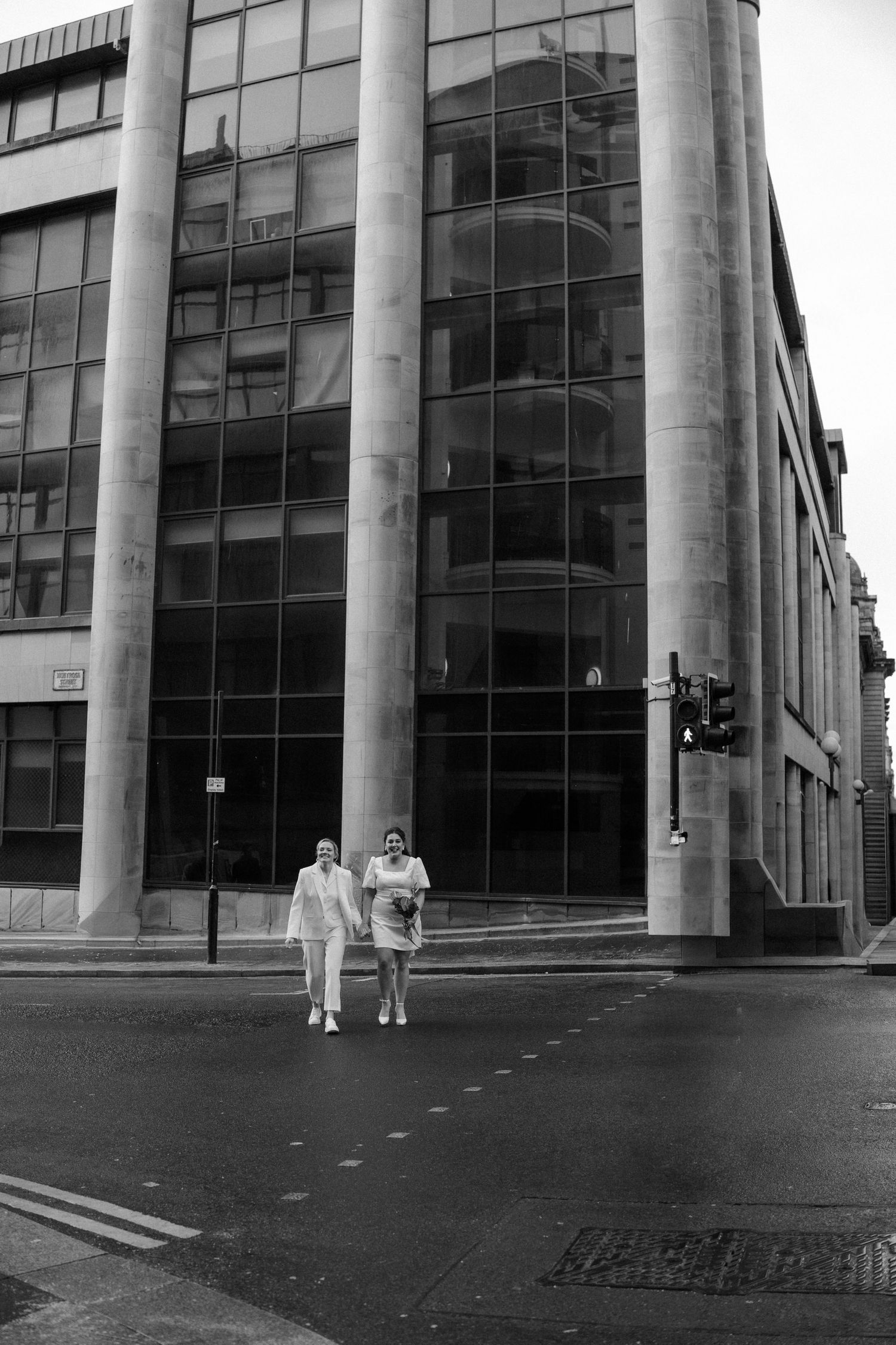 Two brides in white hold hands walking across a city street with a modern glass building behind them.