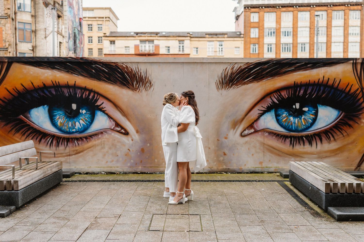 Couple kissing in front of large street art mural of blue eyes on urban wall.