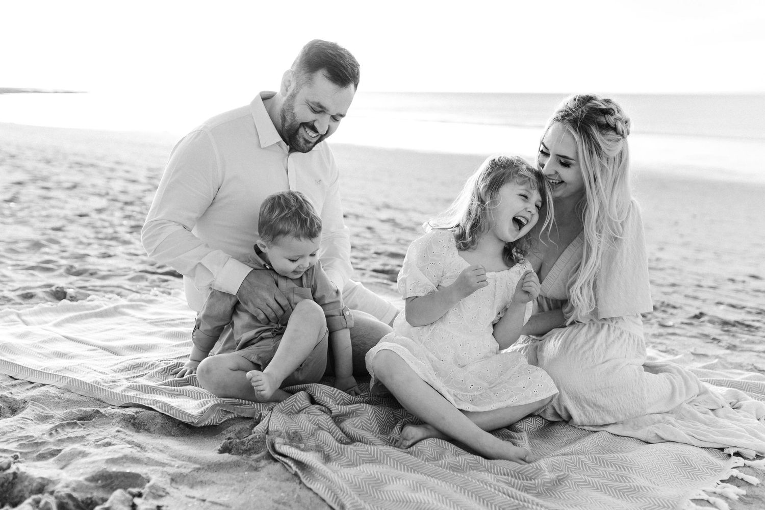 Black and white photo of a happy family of four laughing together on a beach blanket by the ocean.
