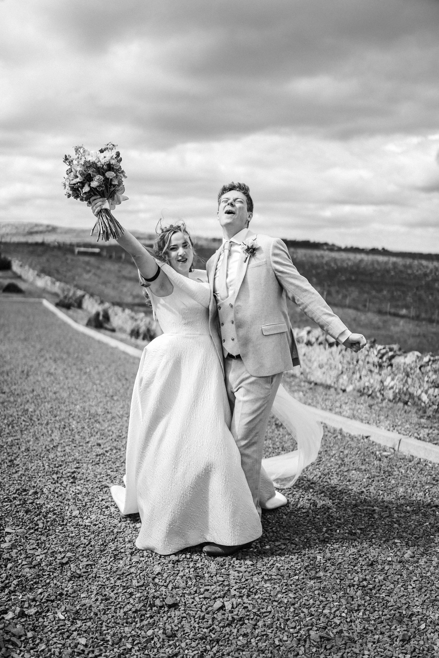 Joyful newlyweds celebrate on a country road, bride raising bouquet high as groom laughs in black and white photo.