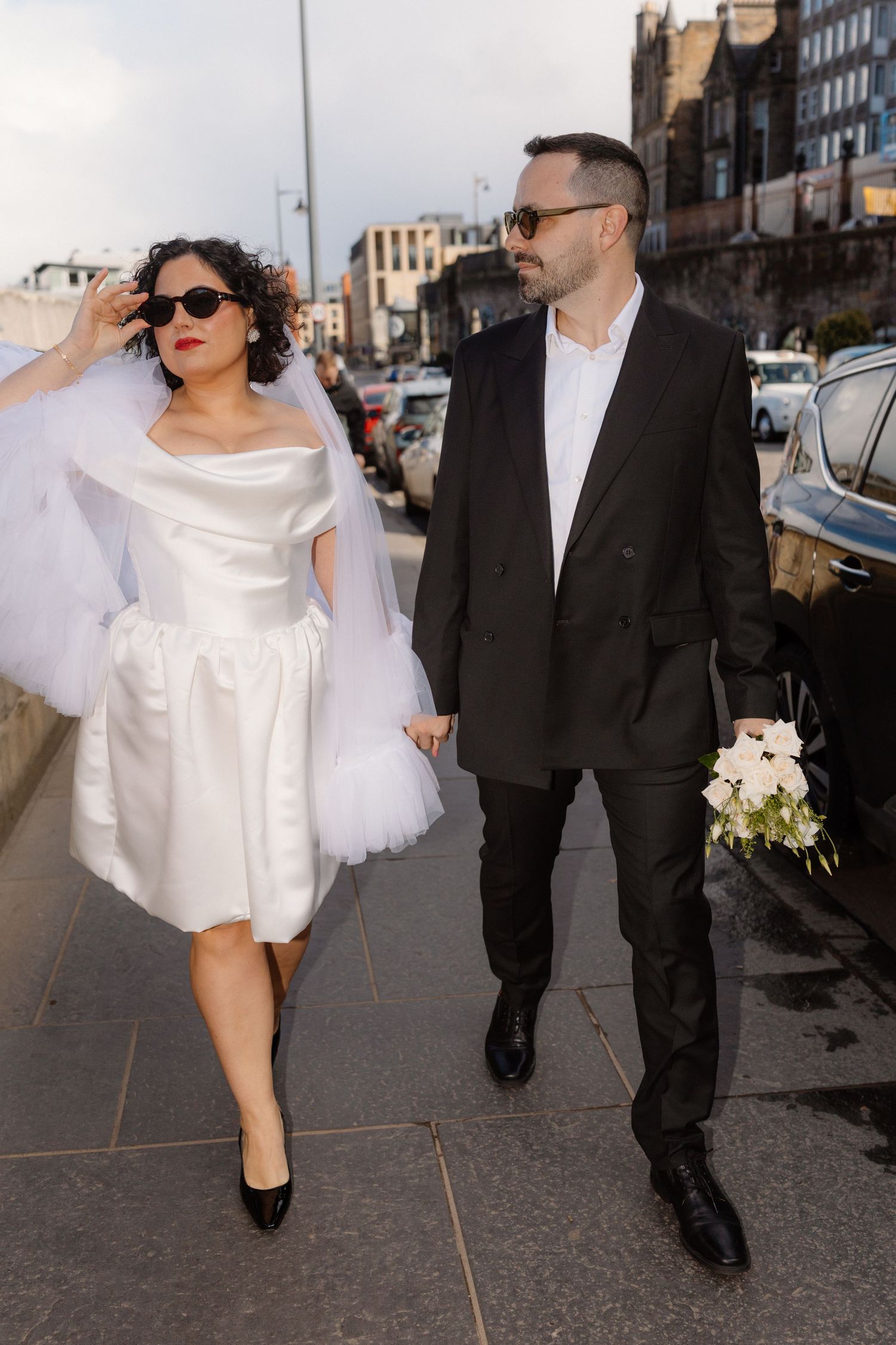 Bride in short white dress and groom in black suit walk hand-in-hand on a city street wearing sunglasses.