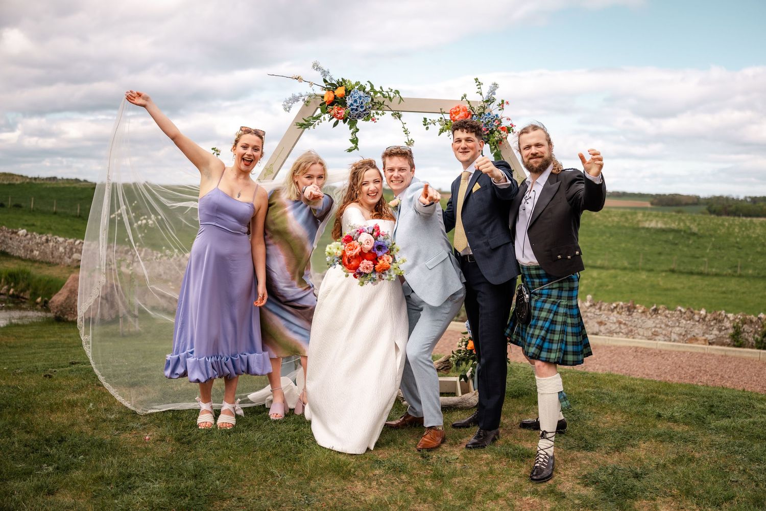 Bride and groom celebrate with wedding party under floral arch in scenic outdoor setting.