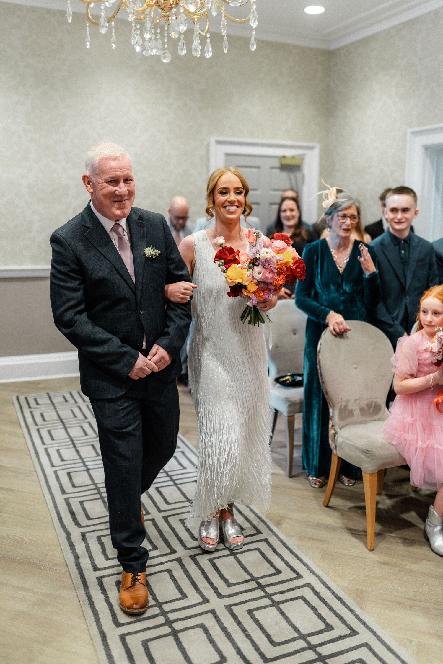Smiling bride walks down aisle with older gentleman holding vibrant bouquet at indoor ceremony.