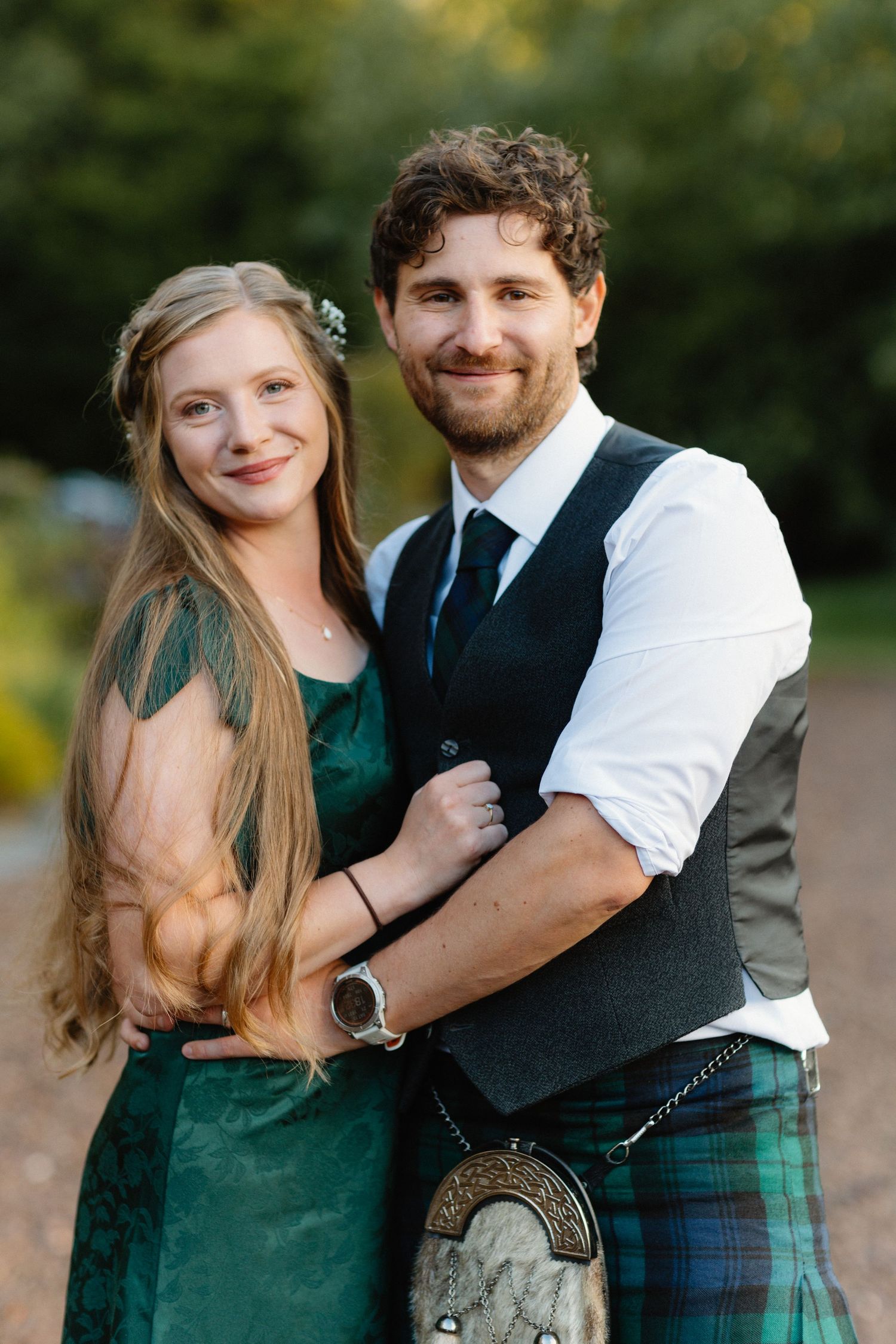 Bride in green dress and groom in tartan kilt embracing outdoors in warm evening light at Scottish wedding.
