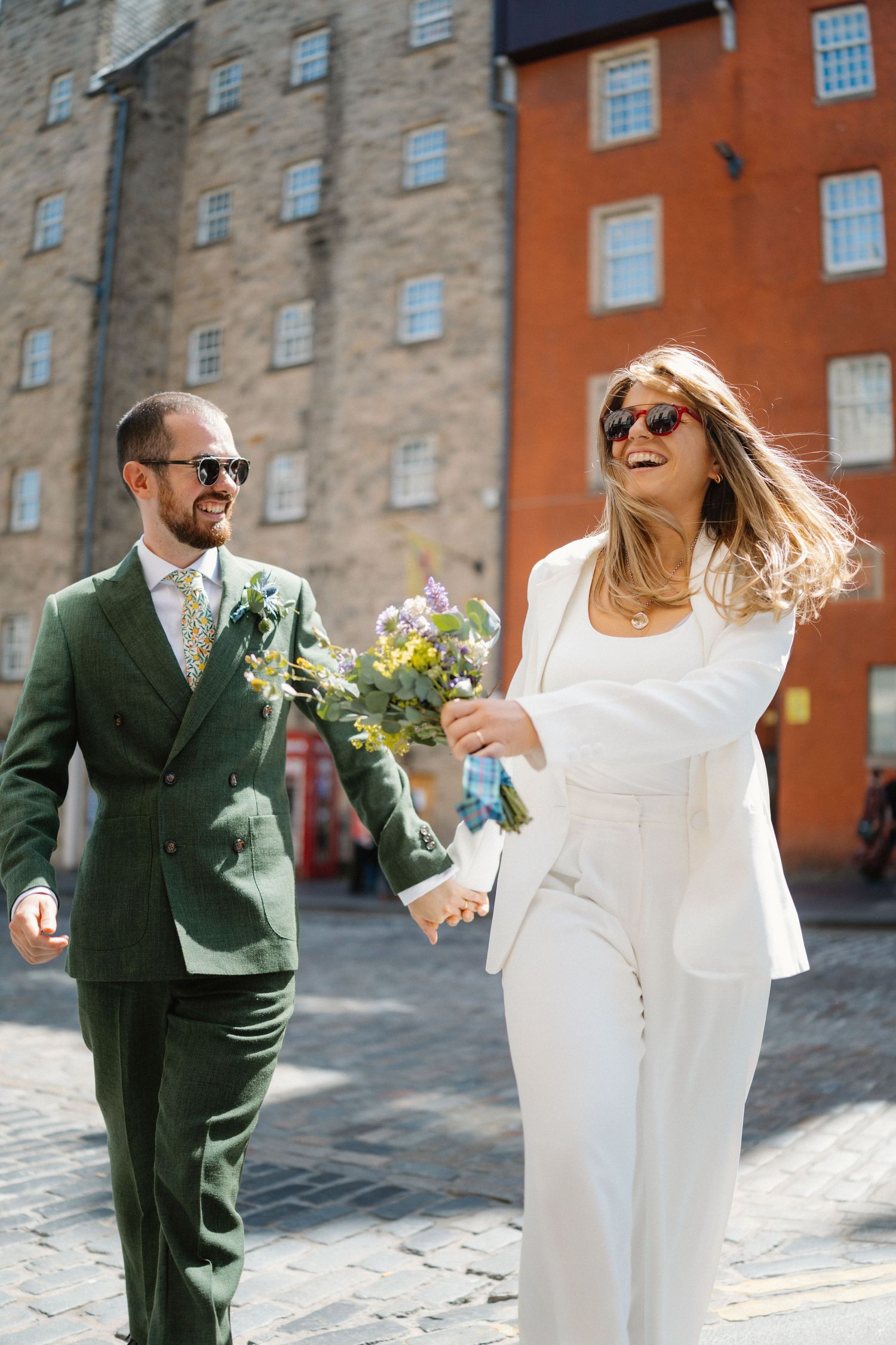 Happy bride in white pantsuit and groom in green suit walk hand-in-hand on cobblestone street holding flowers.