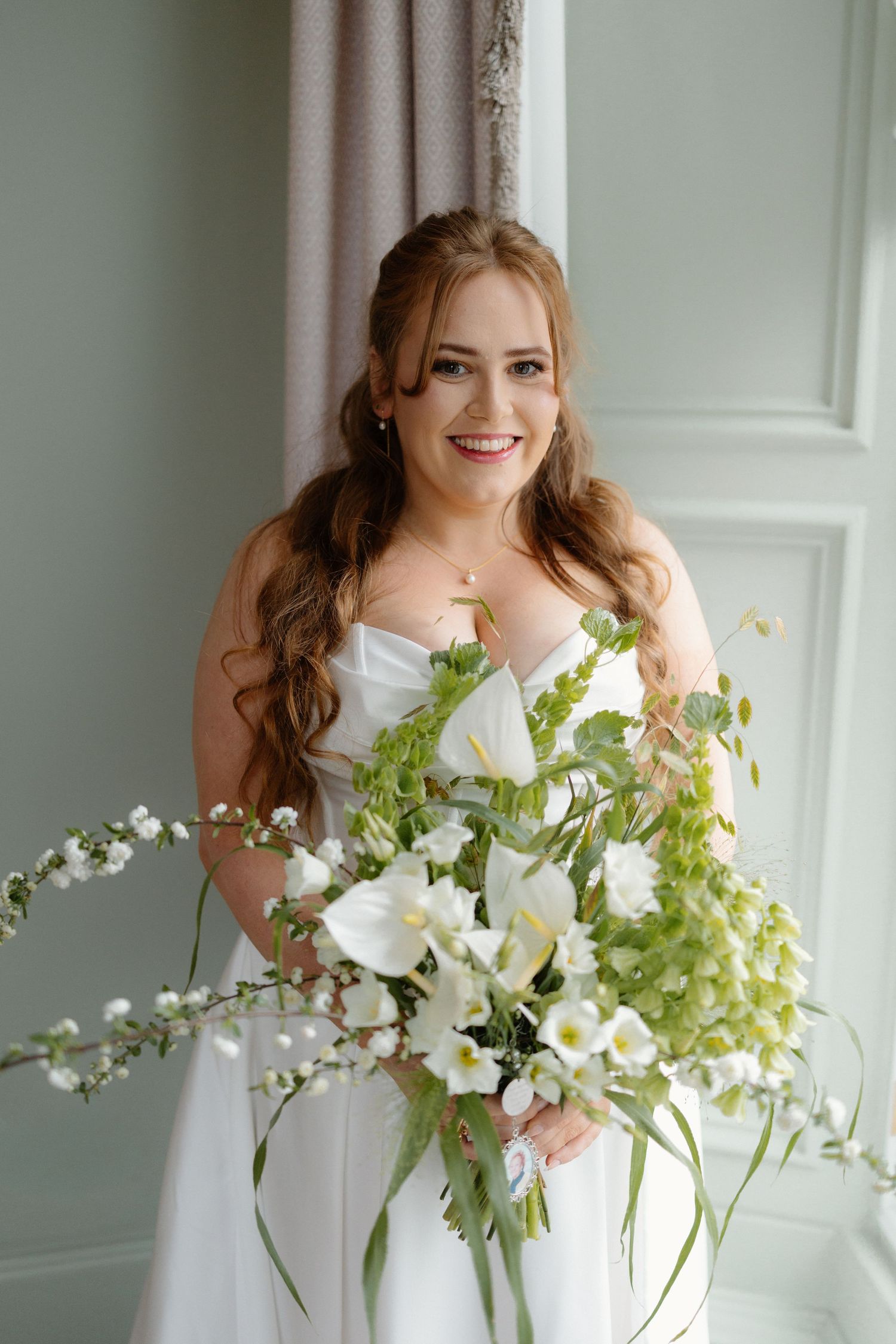 Smiling bride in white strapless gown holding large white and green floral bouquet standing by window.