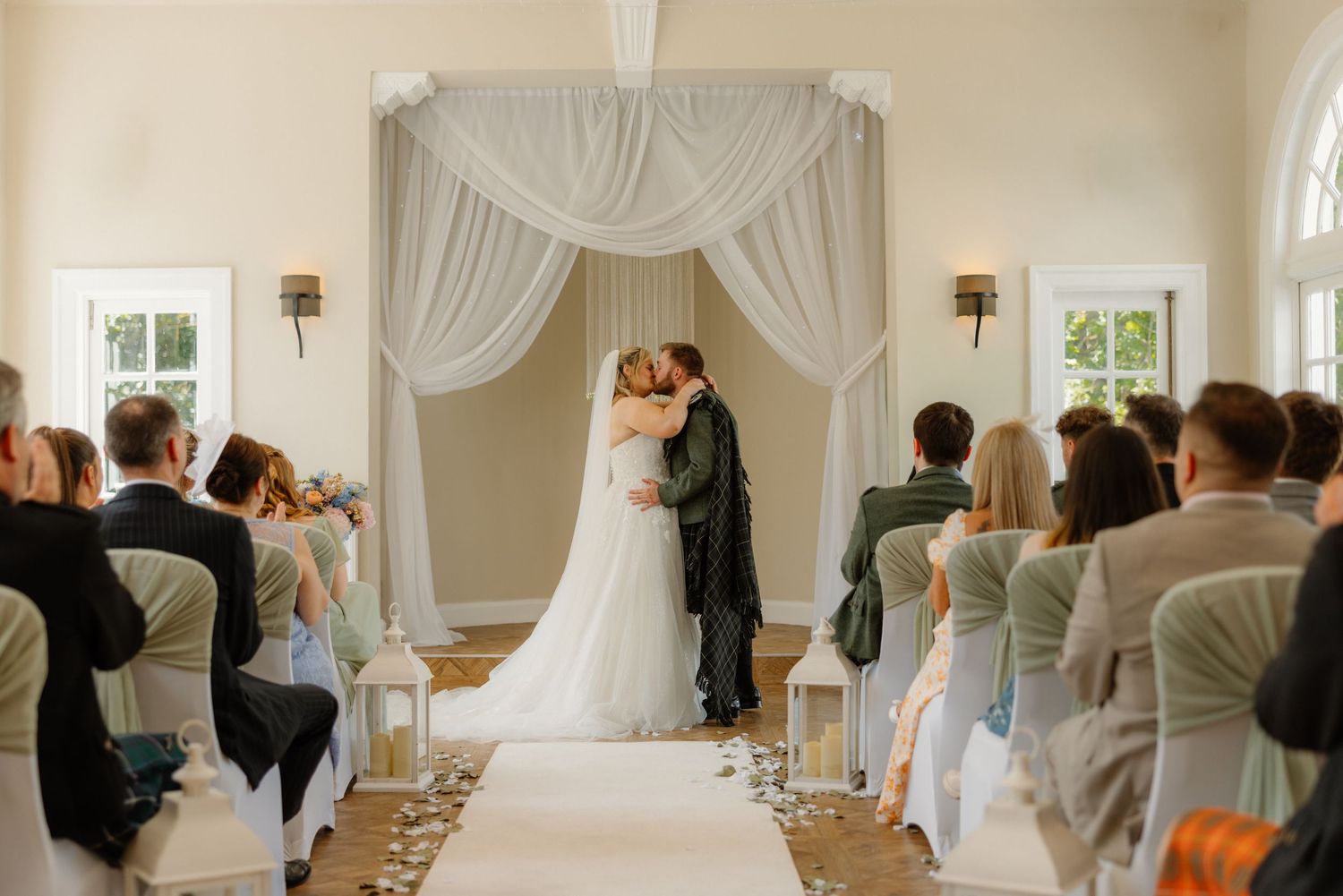 Bride and groom share first kiss at elegant indoor wedding ceremony with white draping and seated guests.