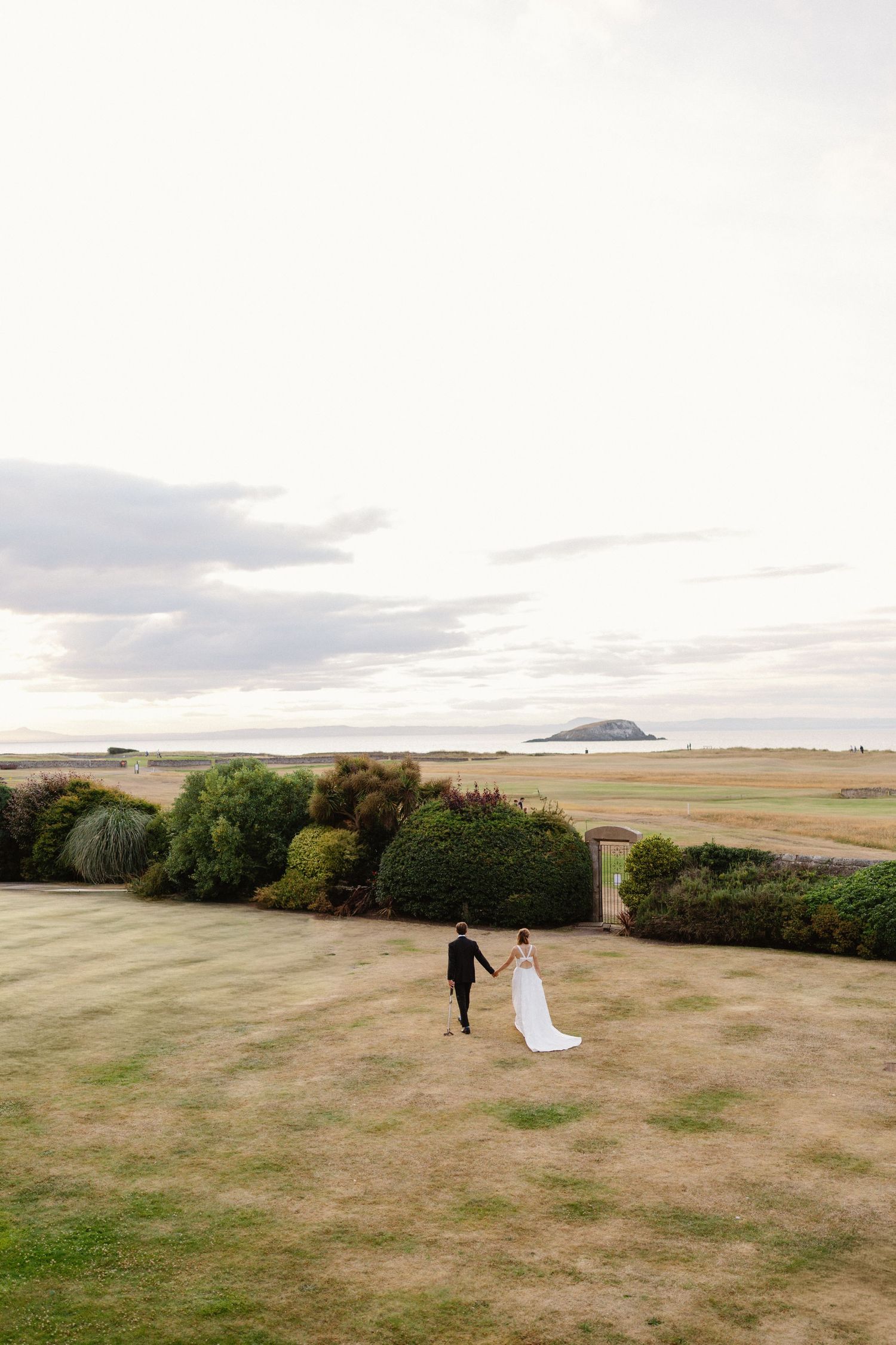Bride and groom walk hand-in-hand across lush green grounds toward a scenic coastal landscape under a cloudy sky.