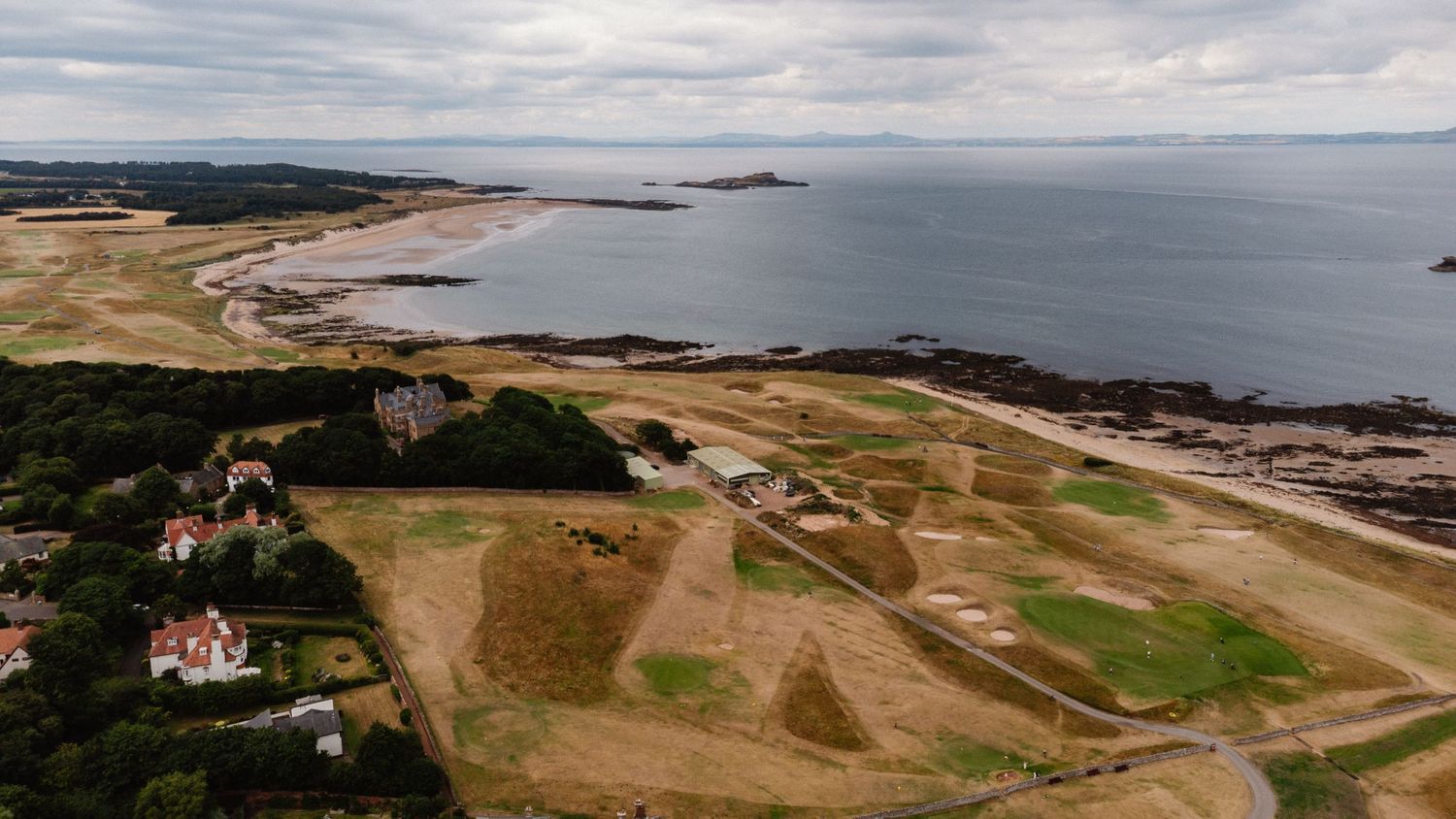 Aerial view of a coastal landscape with sandy beach, rocky shoreline, green fields, and historic ruins under a cloudy sky.
