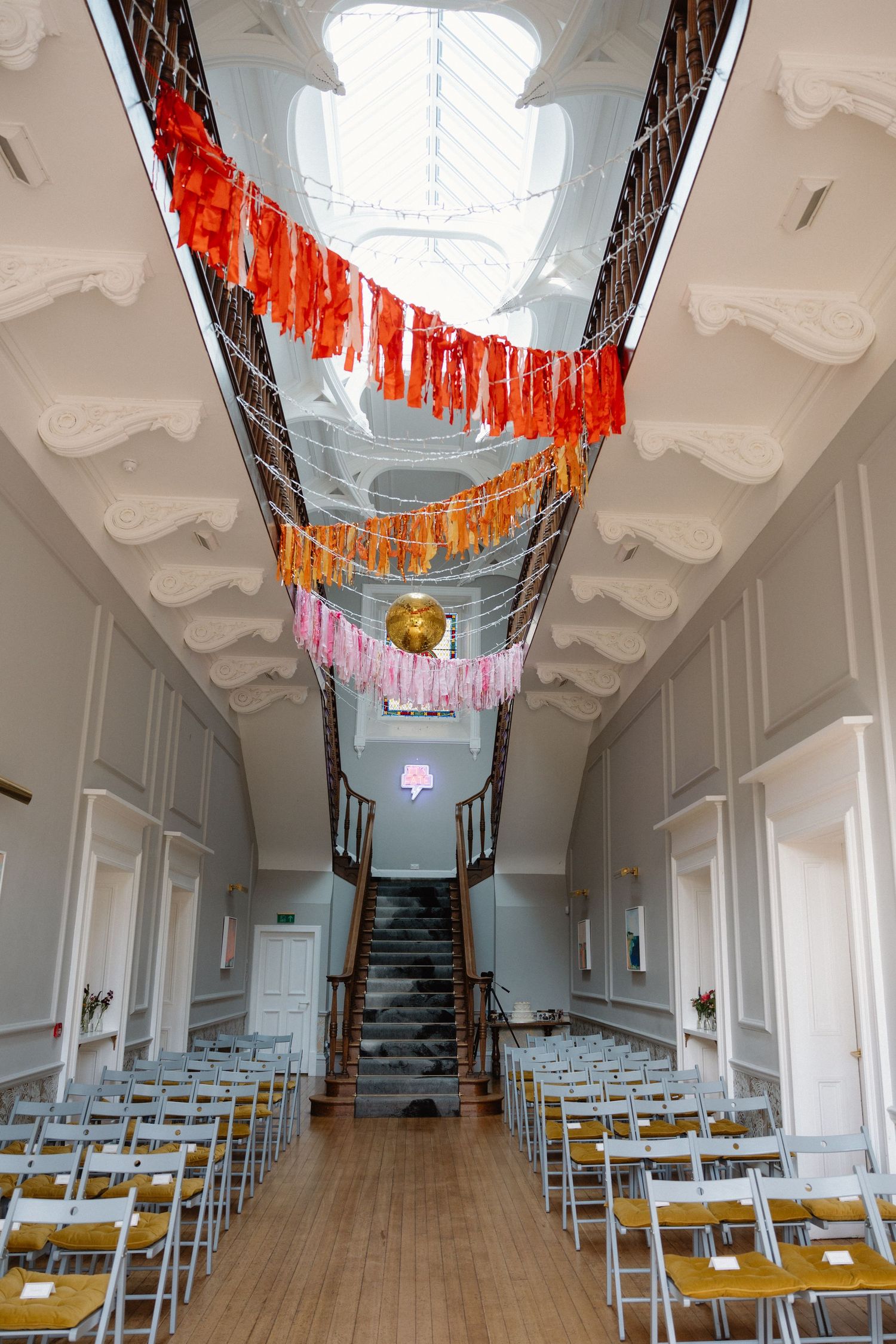 Wedding ceremony space with white walls and ceiling featuring colorful hanging decorations above rows of gold chairs.