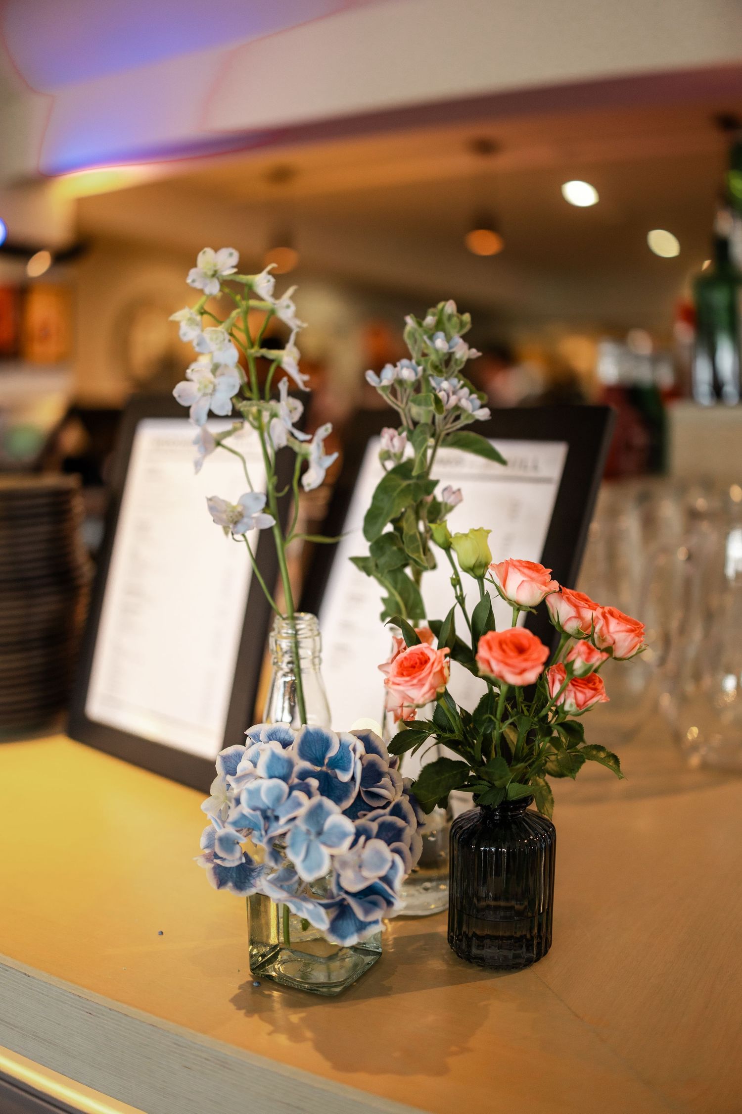Elegant floral centerpieces with coral roses and blue hydrangeas in small vases on reception tables.