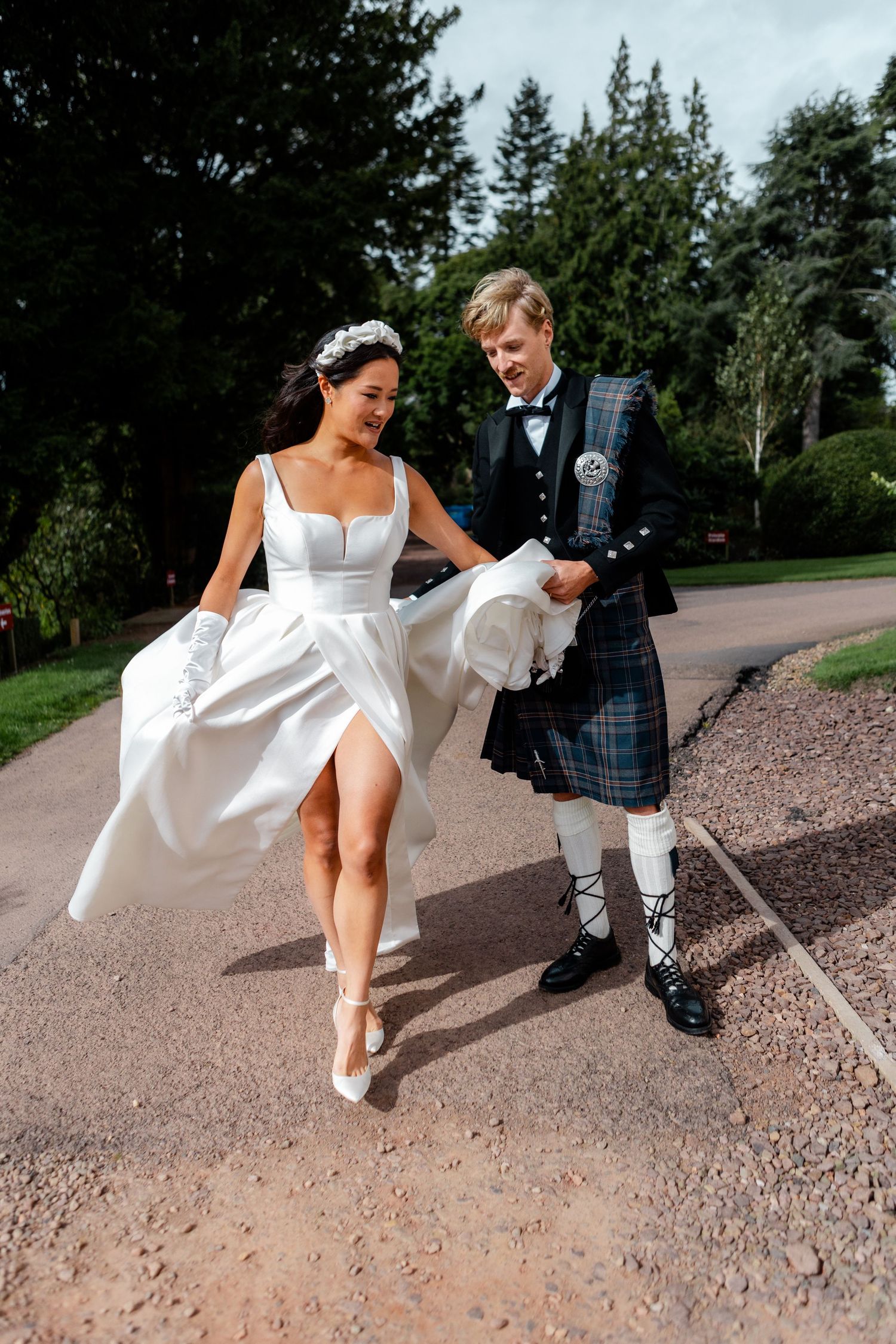 A playful wedding photo with the bride twirling her dress next to a groom in traditional Scottish attire.