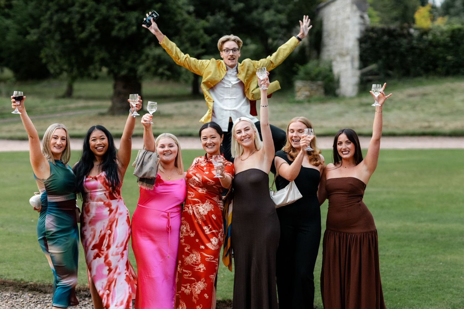 Wedding party members in colorful dresses celebrate joyfully on a grassy lawn.