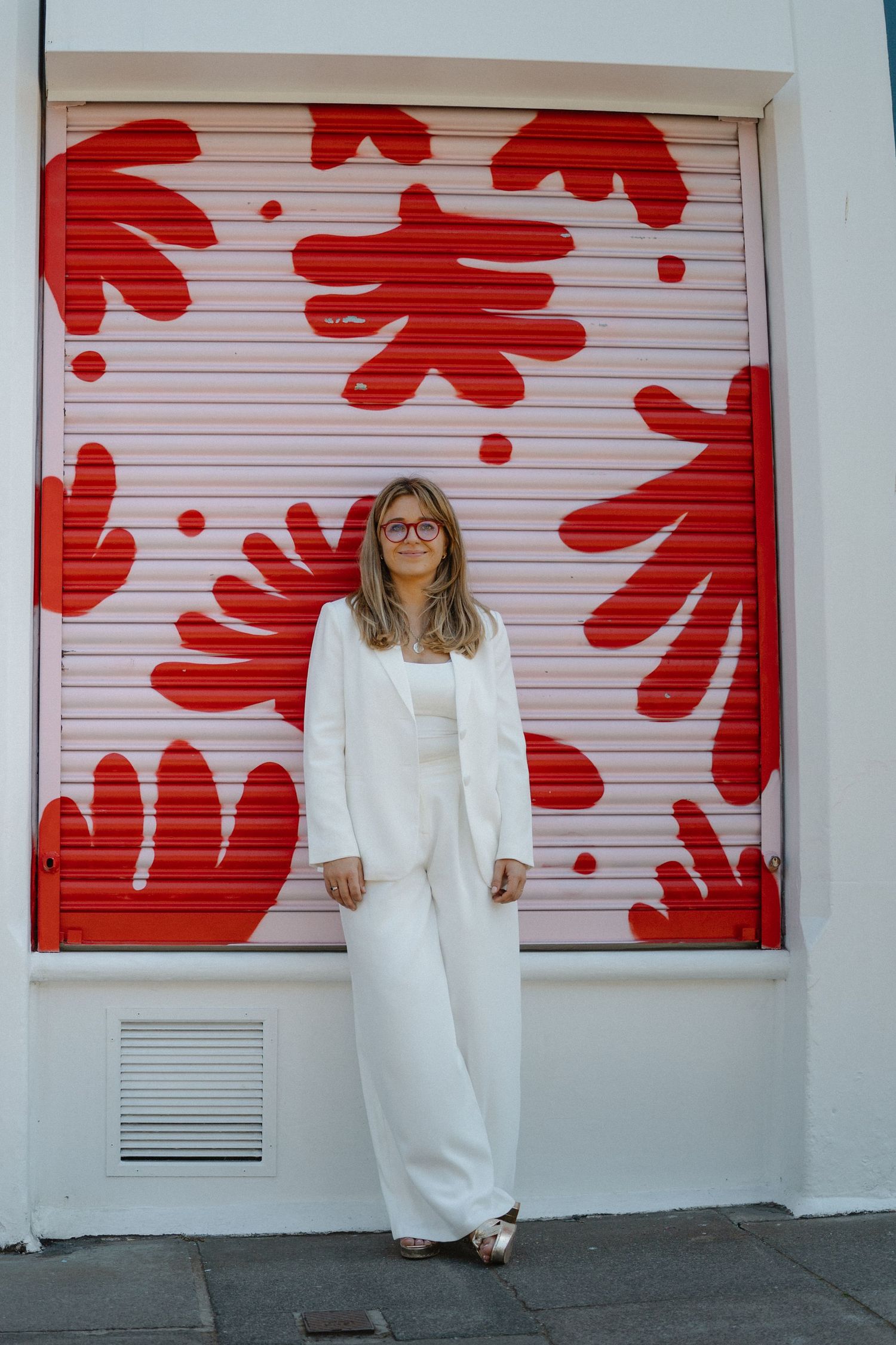 A person in white clothing poses against a wall with bold red floral patterns.