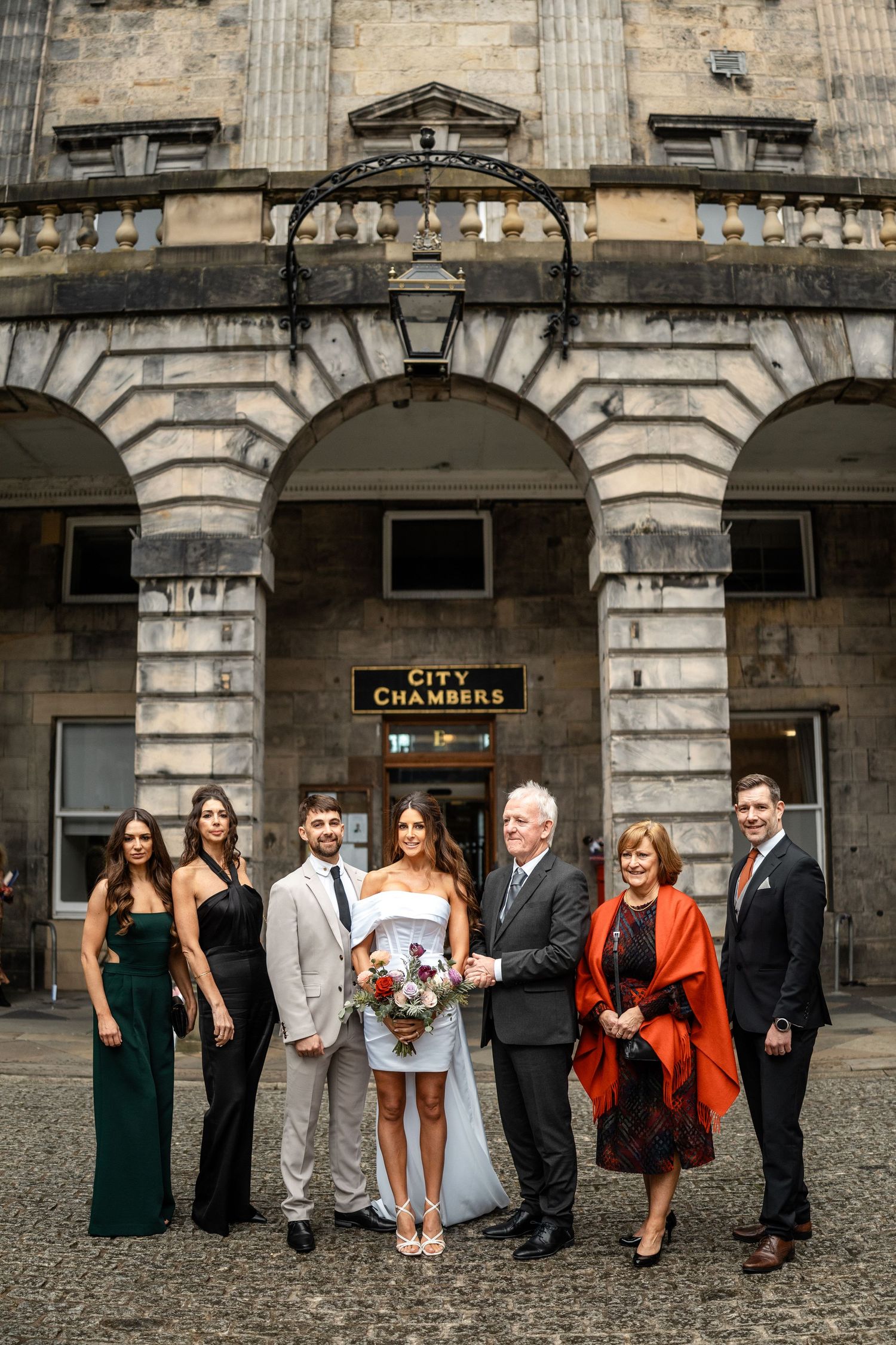 Wedding party poses outside historic stone building with arched architecture.