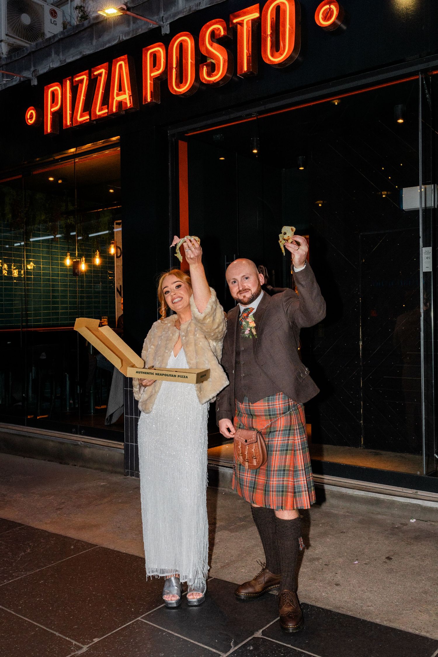 Happy couple celebrating outside Pizza Posto with neon sign glowing above.