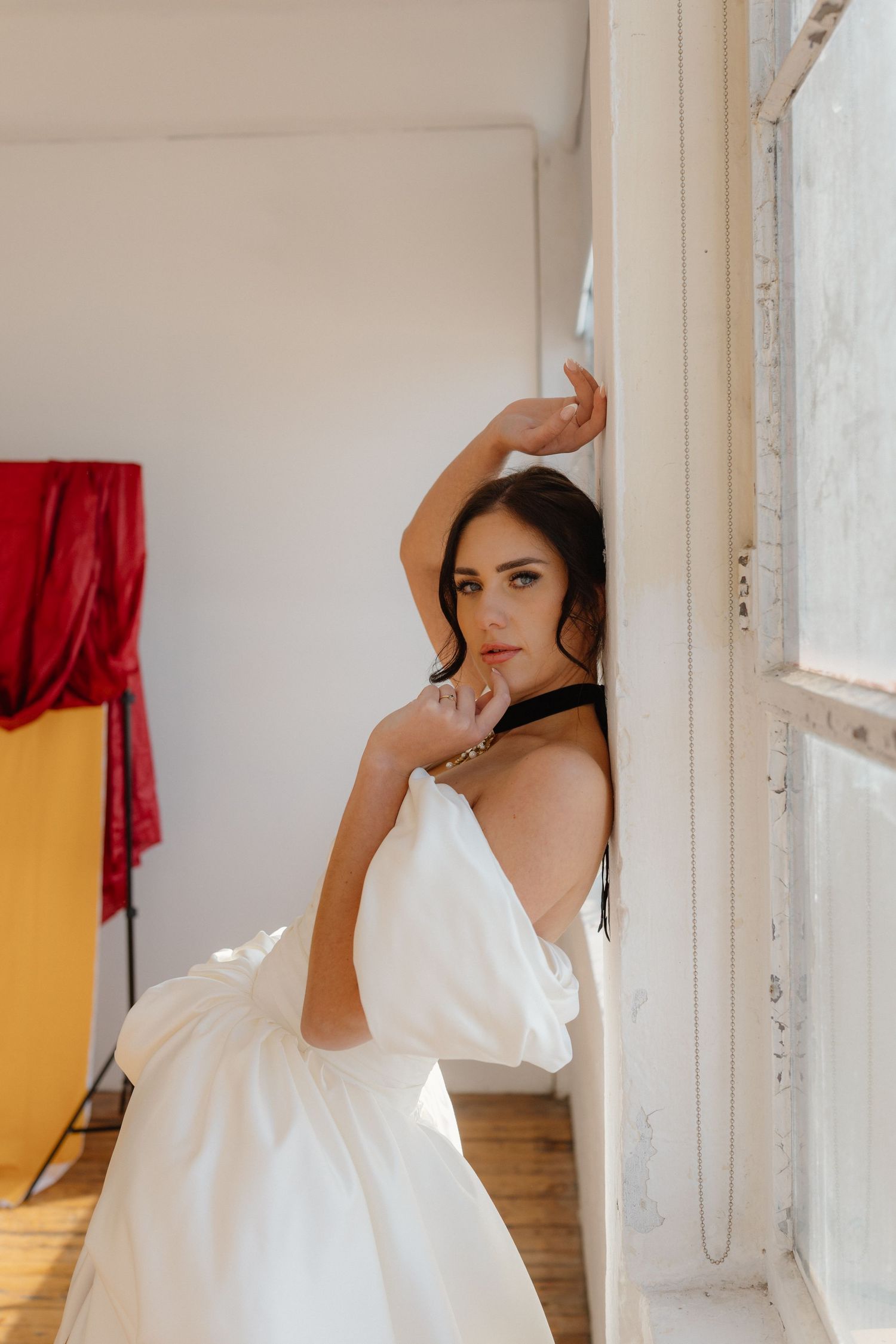 Woman in white gown leans against window frame with arm raised, wearing black choker, in bright studio space.