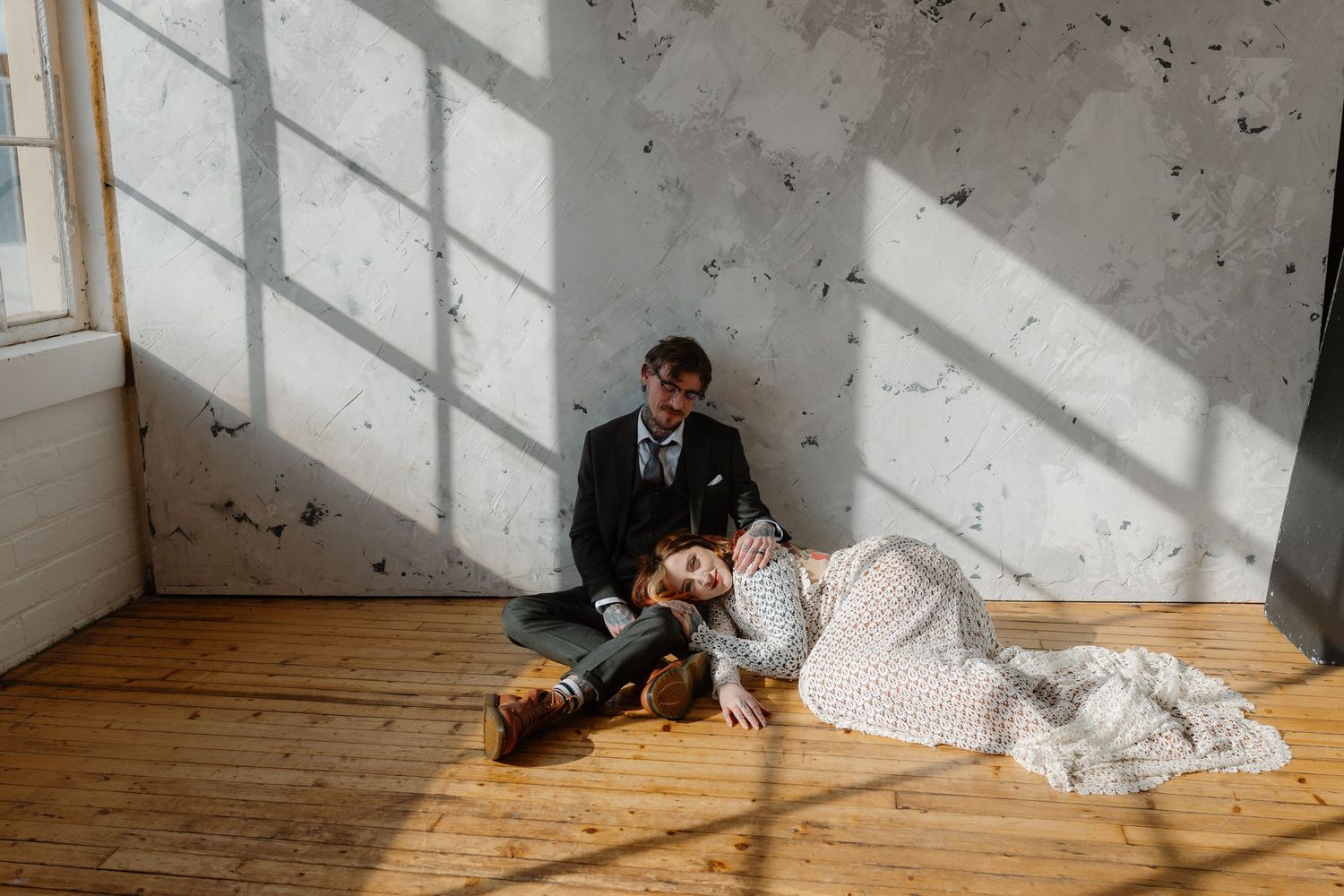 Bride and groom sitting on wooden floor in sunlit room, sharing a romantic moment in wedding attire.