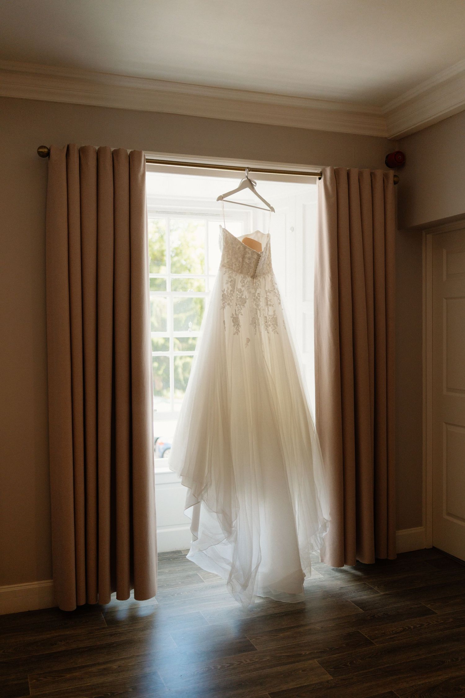 Elegant strapless lace wedding gown hanging in a sunlit window with flowing tulle skirt on display.