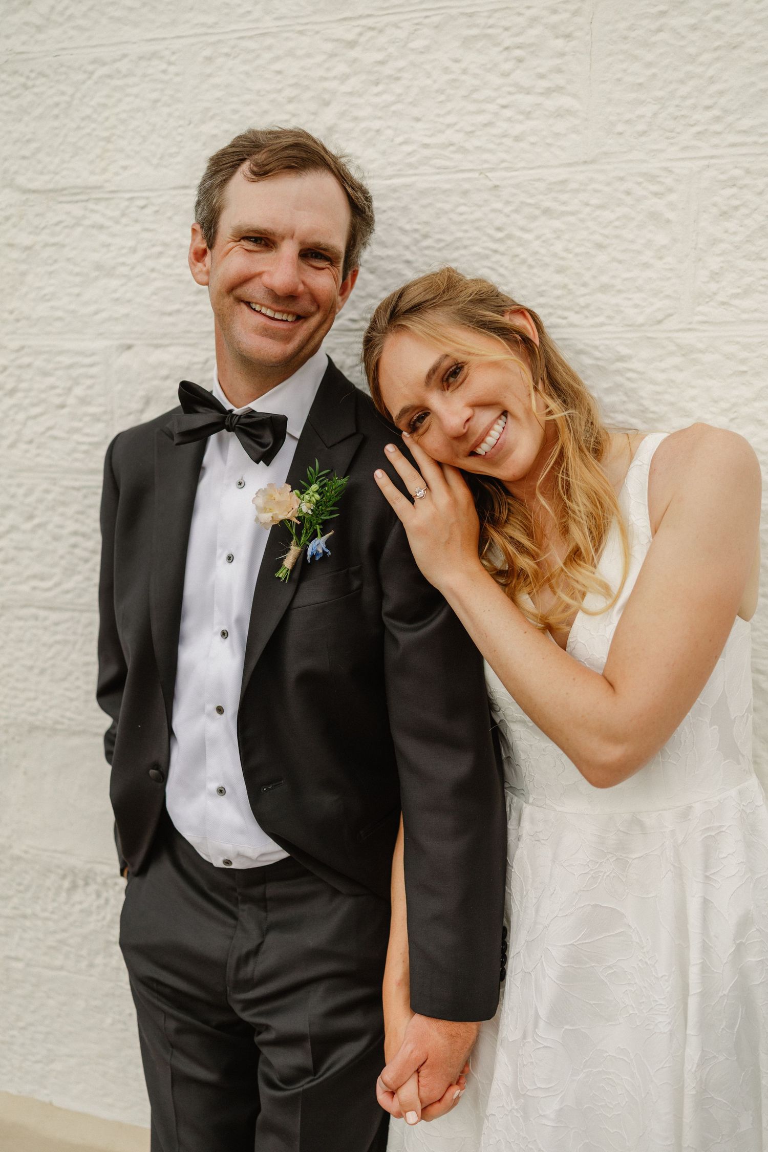 Smiling bride and groom in black tuxedo with boutonniere posing against a white textured wall.