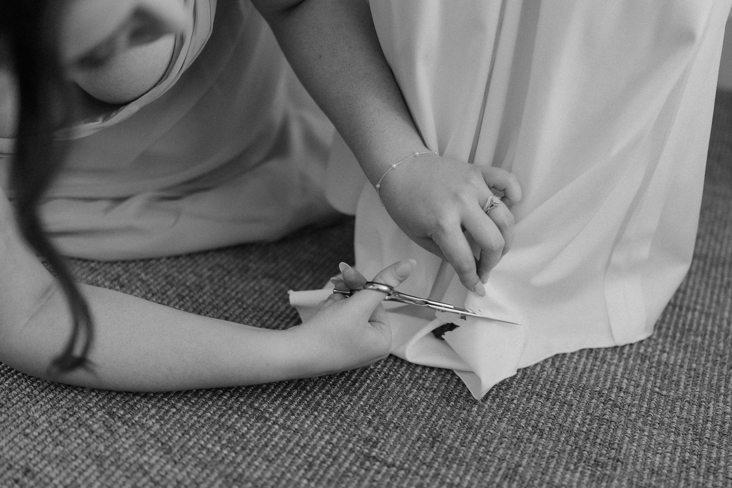Bridesmaid kneeling on carpet adjusting the hem of a white wedding dress in black and white photo.
