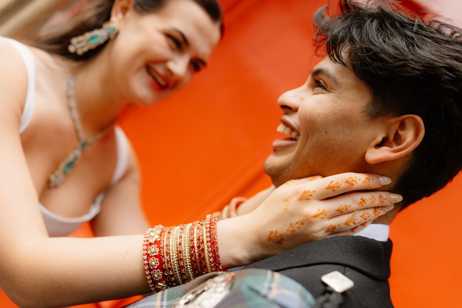 A joyful couple with henna hands and colorful bangles share a smile against a vibrant orange background.