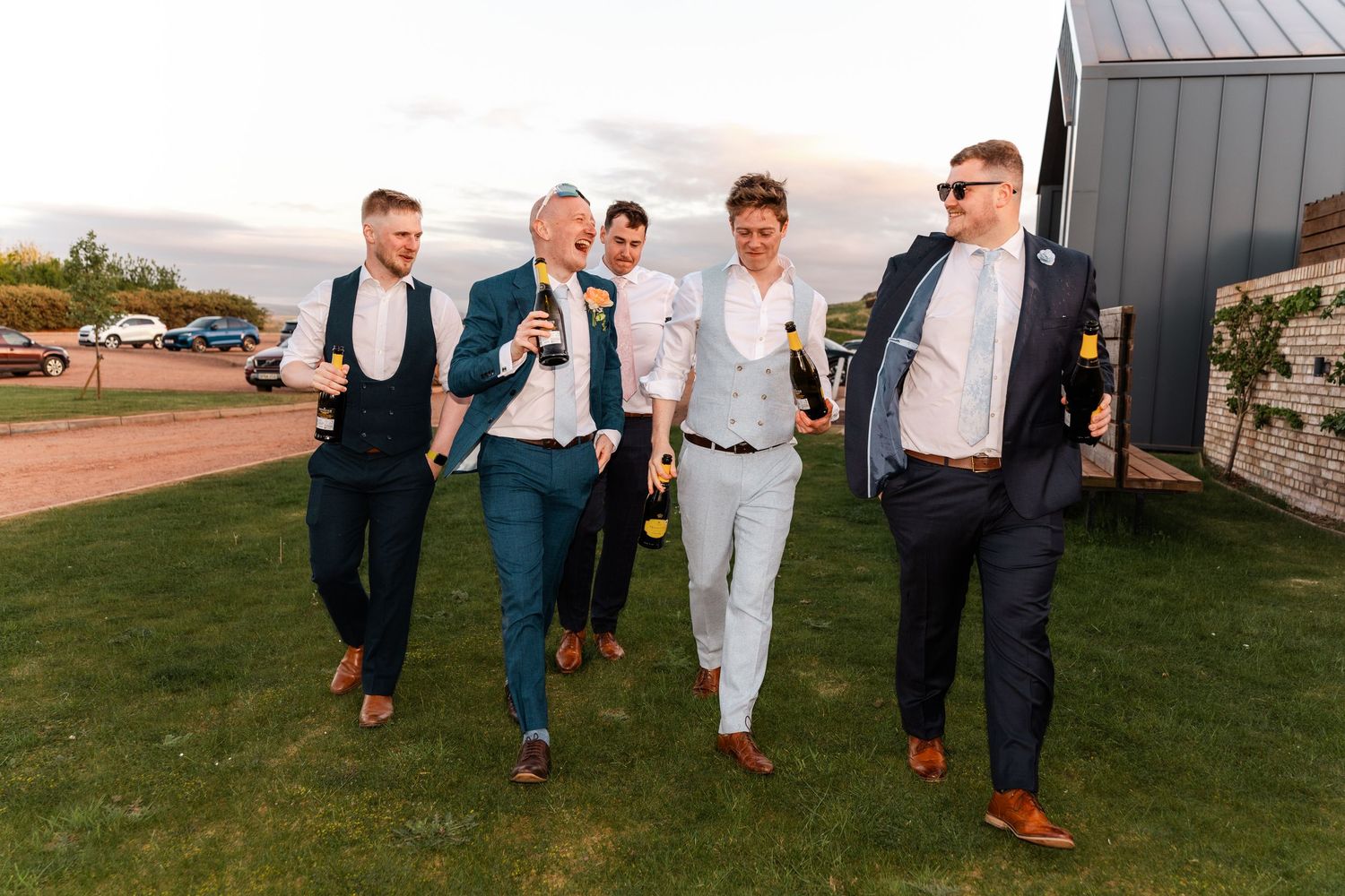 Groom and groomsmen in suits walk together holding drinks at outdoor wedding venue.