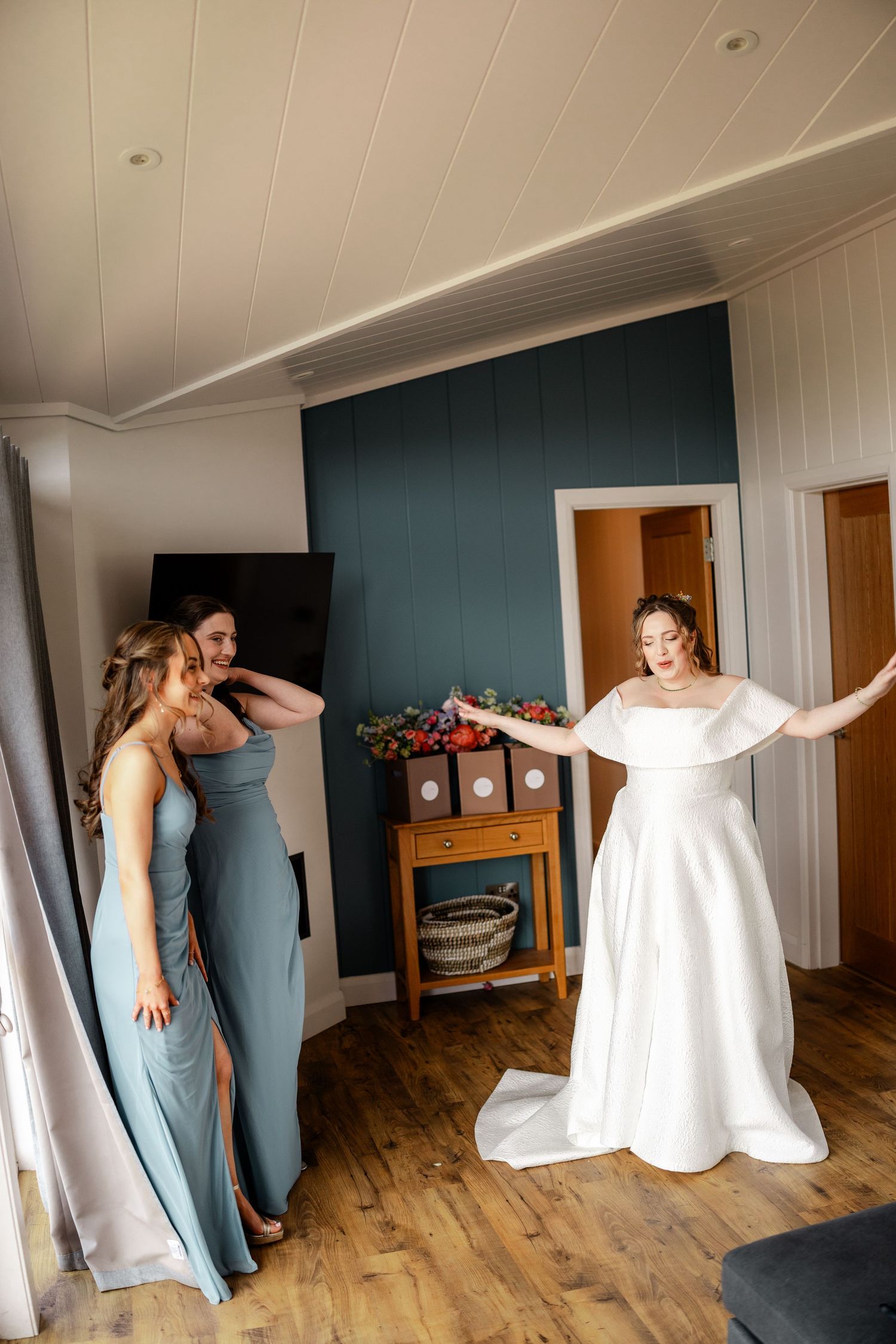 Bride in white gown reveals dress to bridesmaids in blue dresses during wedding preparation.