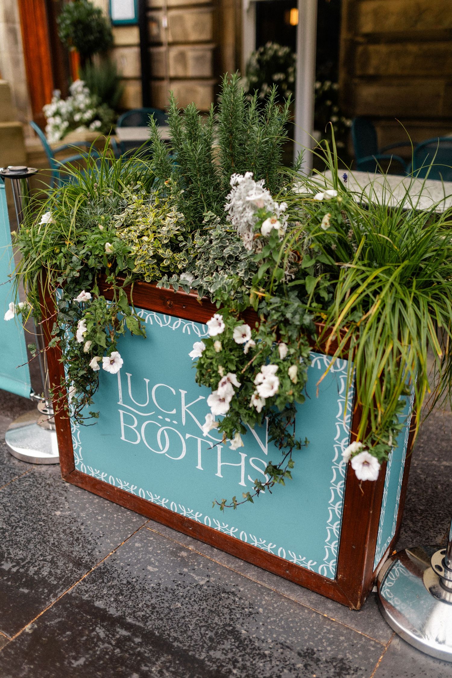 Decorative teal Lucky Booths planter box filled with lush green plants and white flowers outdoors.