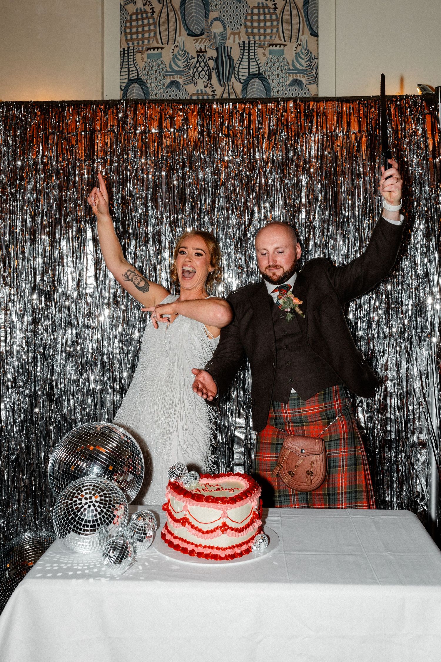 Bride and groom celebrating by a red wedding cake with disco balls and silver tinsel backdrop.