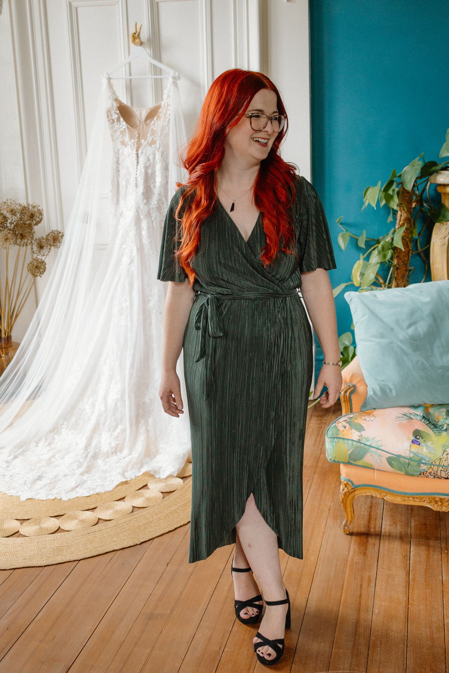 Woman with vibrant red hair in elegant green wrap dress standing near bridal gown in decorated room.