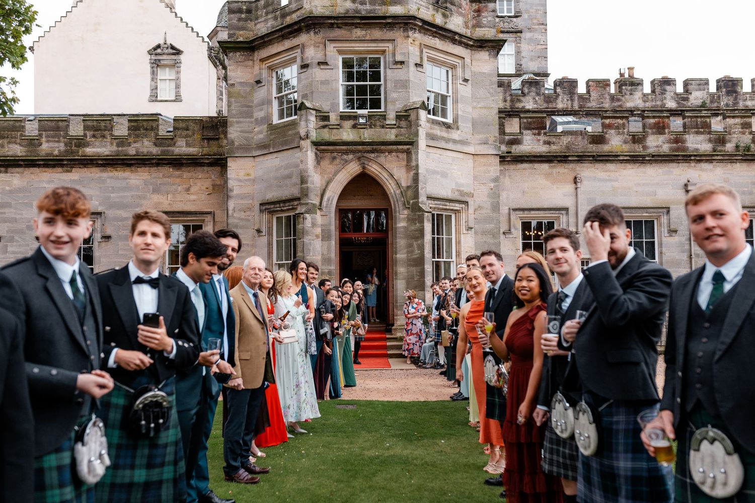 Wedding guests in kilts and formal wear lining a red carpet outside a historic Scottish castle venue.