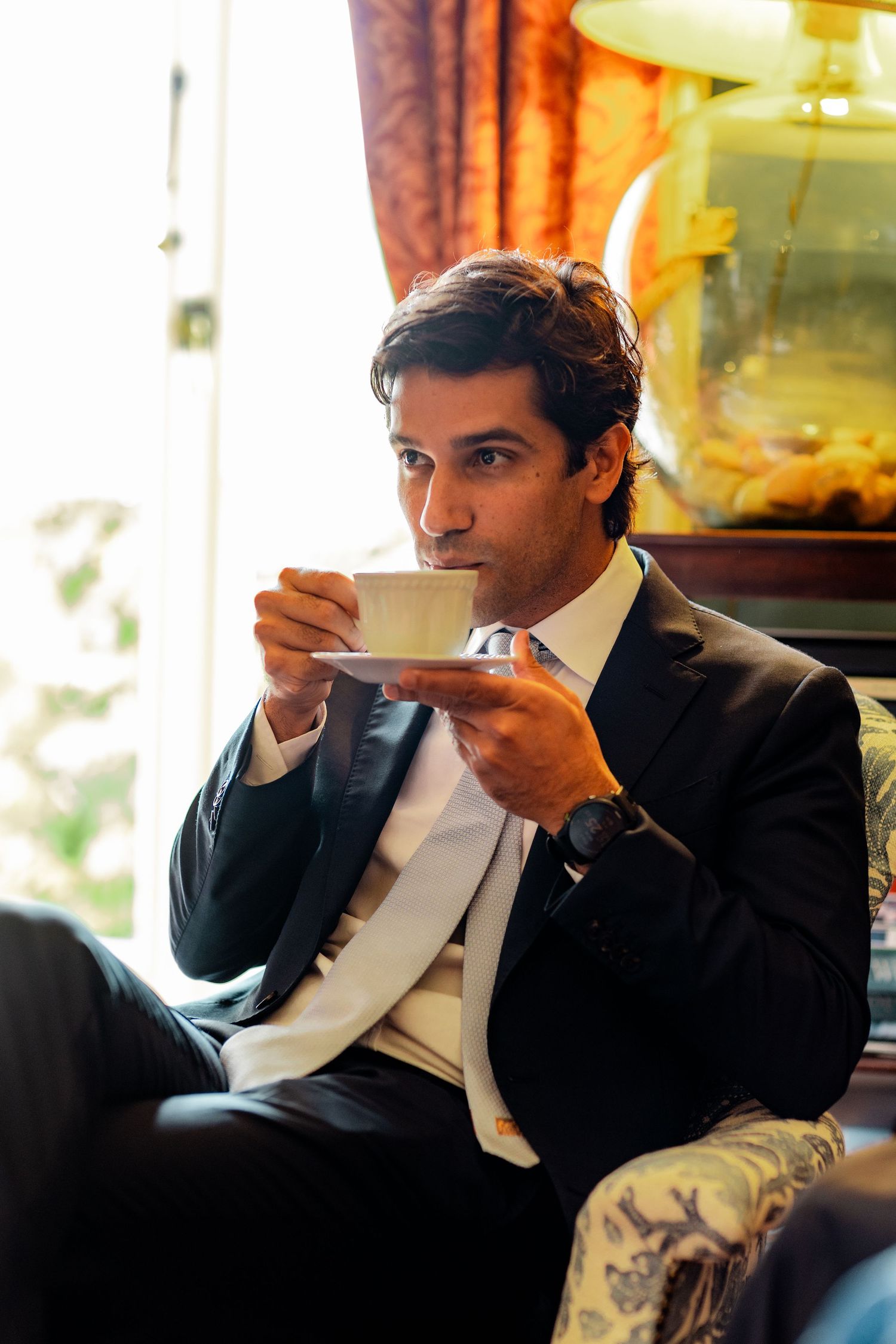 Elegant man in black suit drinking from a teacup while seated in a luxury interior.