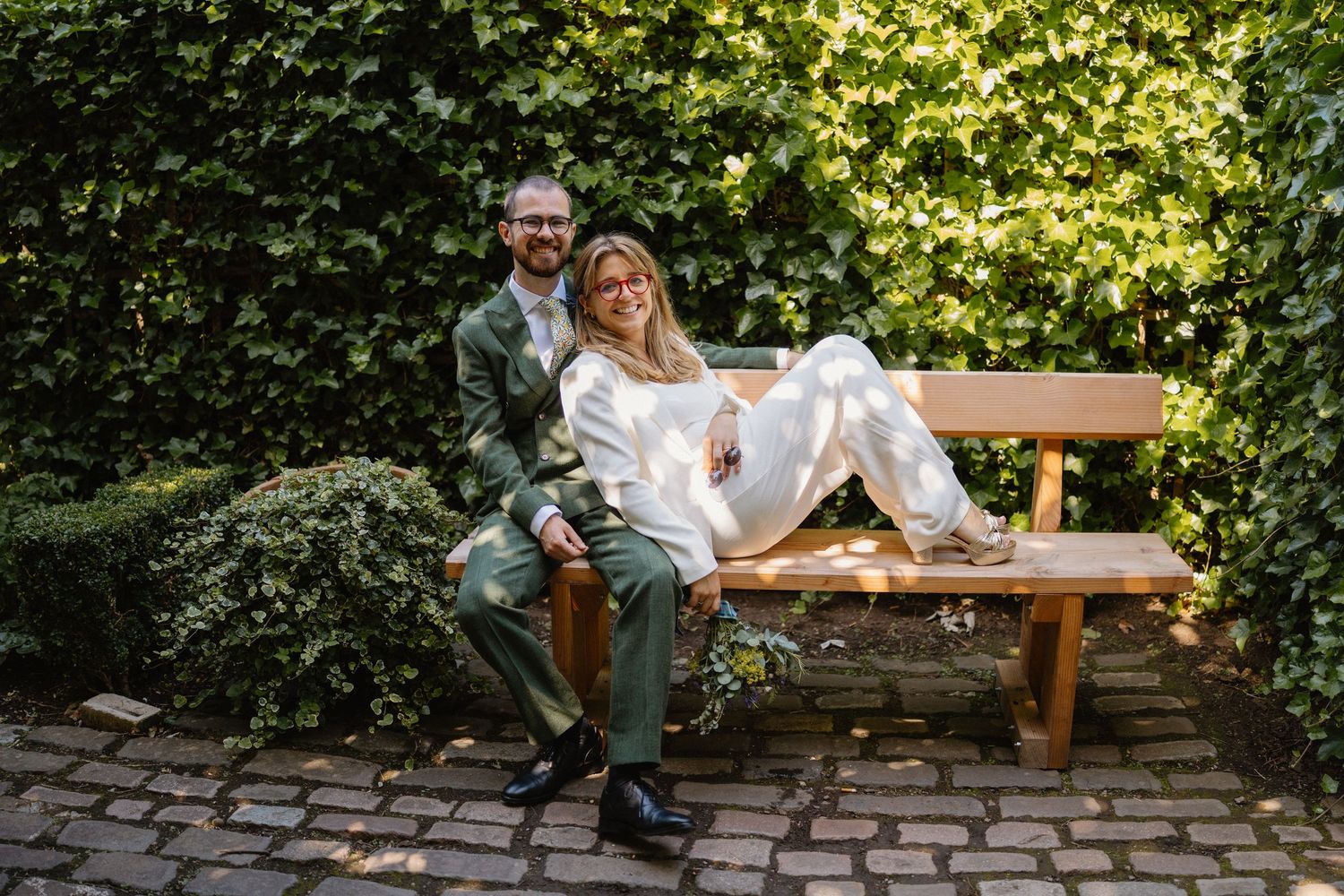 Bride and groom sit on wooden garden bench surrounded by lush green ivy walls after wedding.