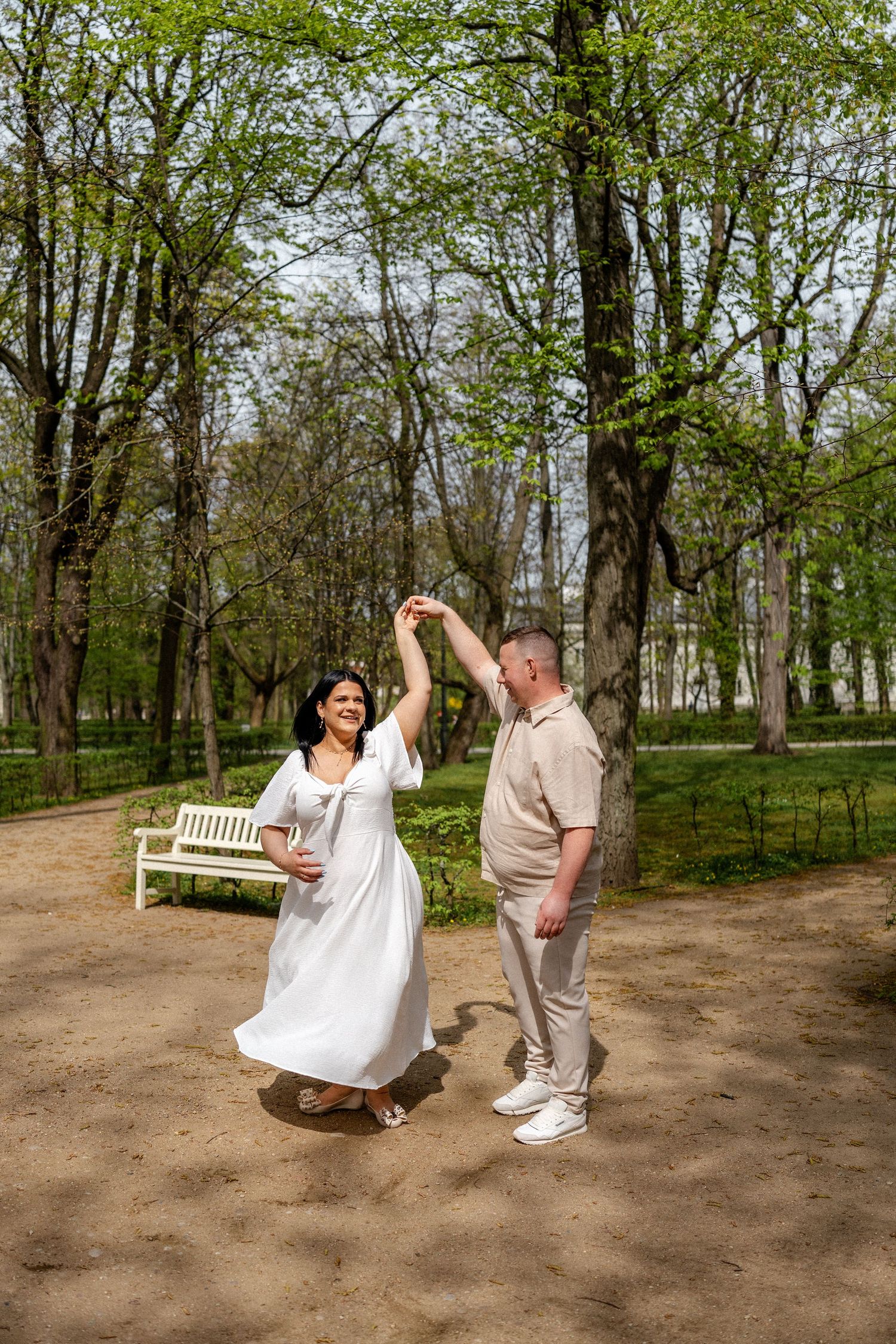 A couple playfully poses together on a dirt path surrounded by trees in a park setting.