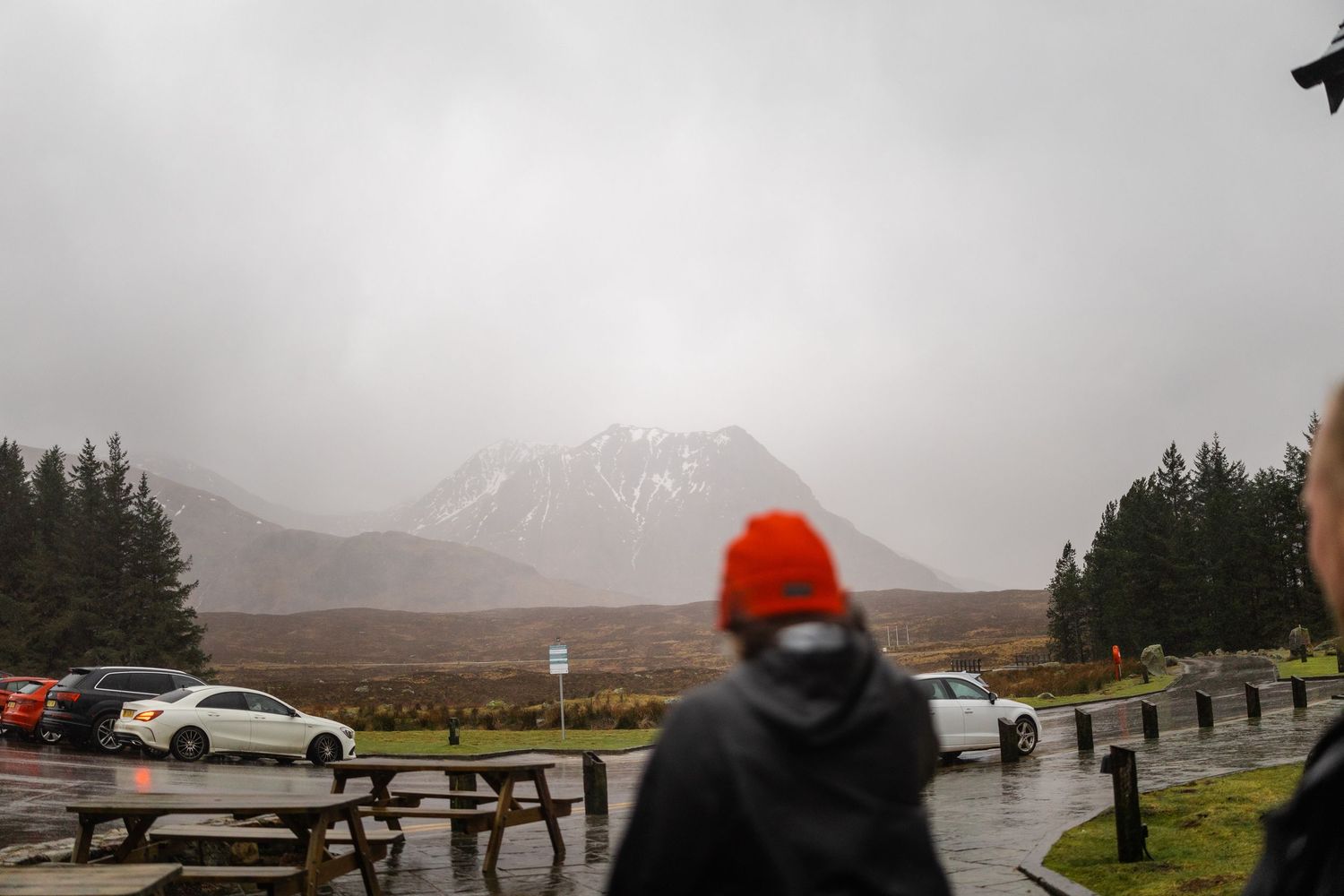 Person in orange beanie looking at snow-capped mountain from parking area.