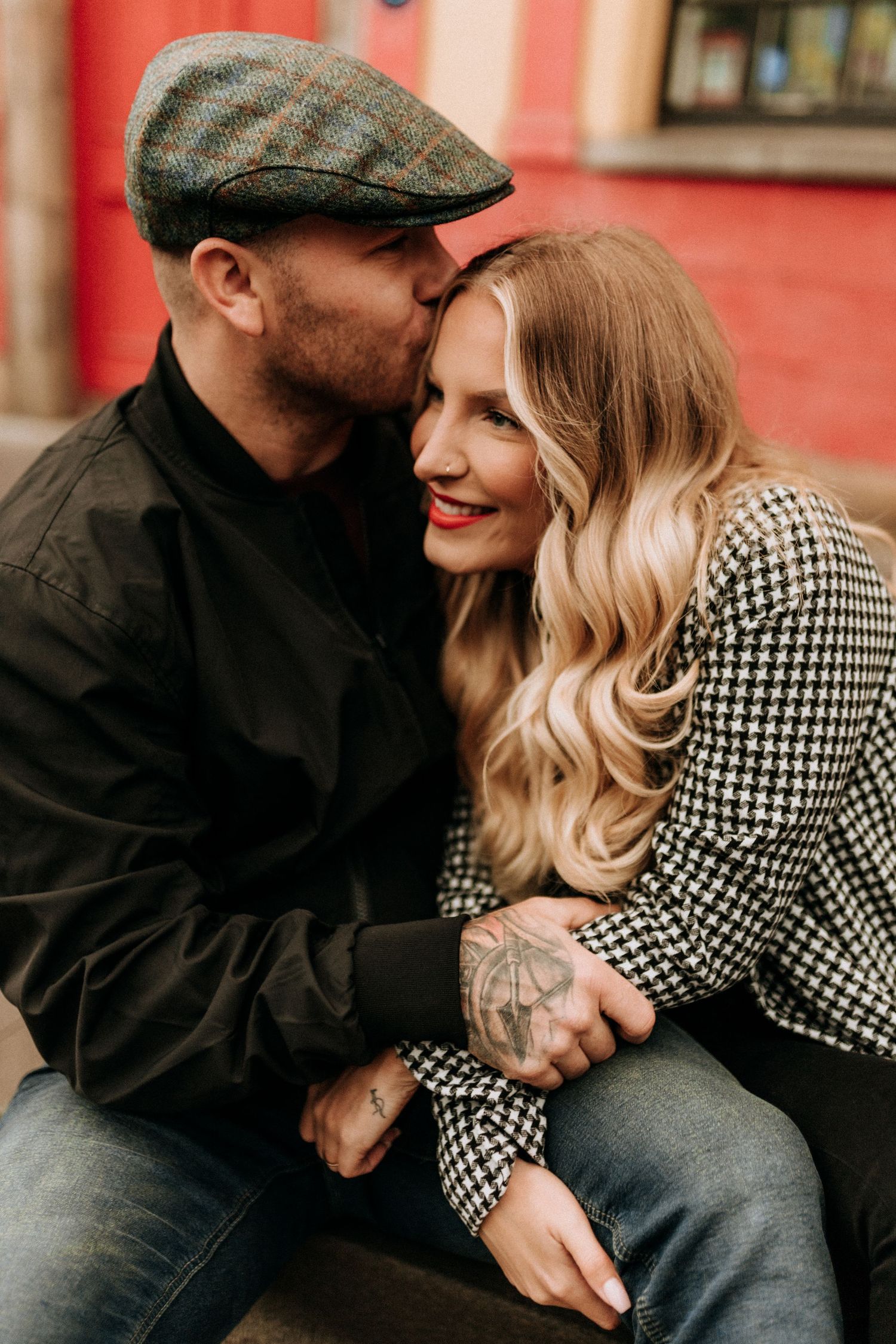 Intimate couple in black and houndstooth pattern sharing tender moments against a red wall background.