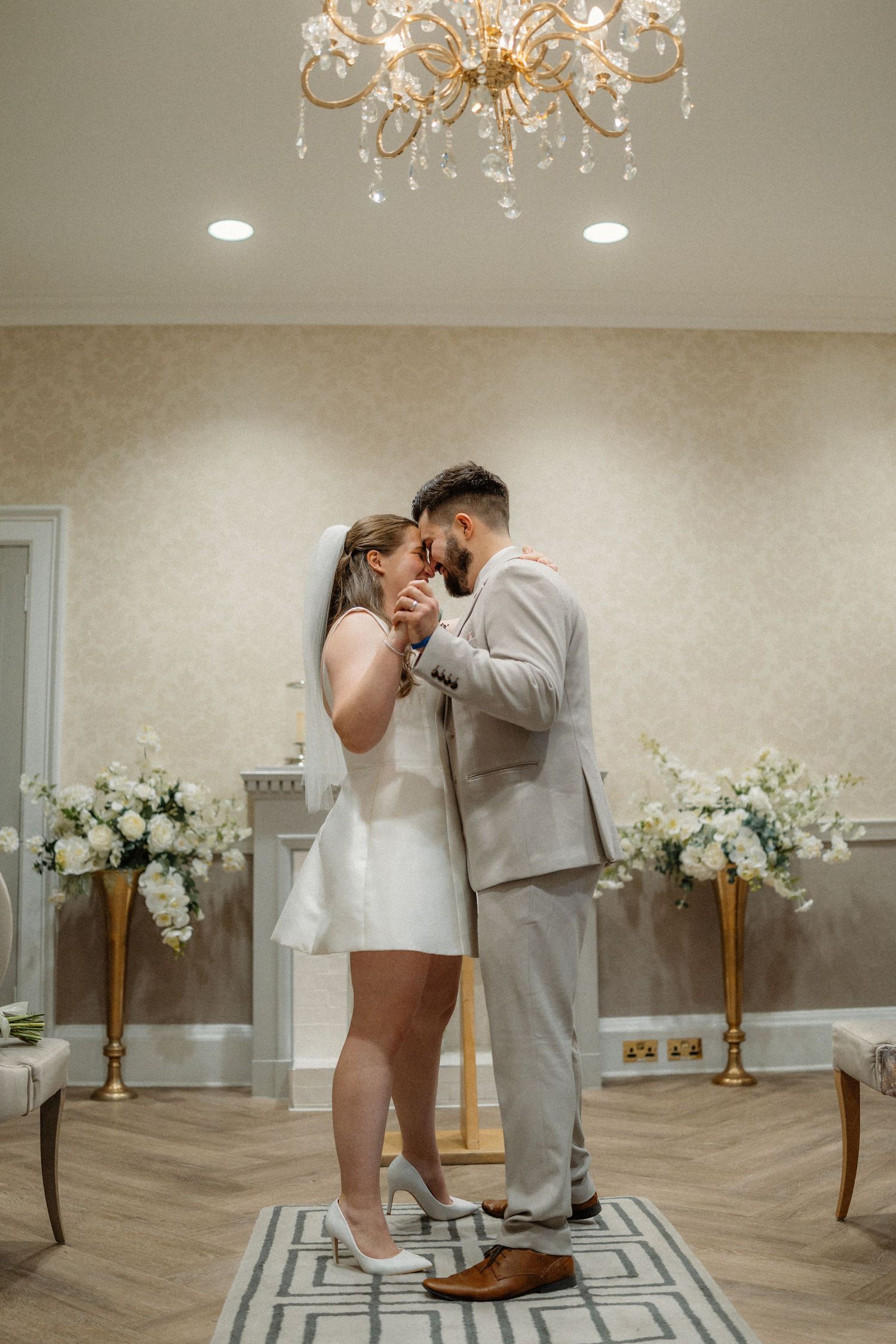 A couple shares a romantic dance moment under a crystal chandelier in an elegant white room with floral decor.
