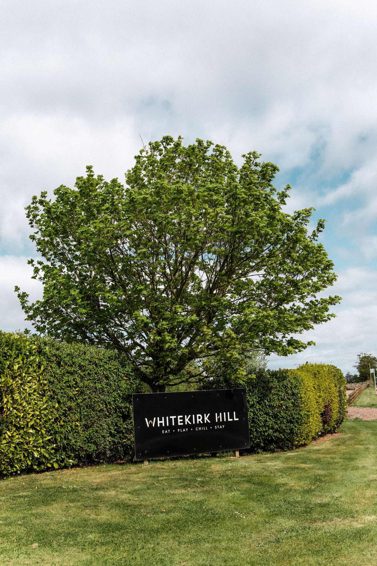 A panoramic view of Whitekirk Hill with lush green trees and a sign against a cloudy sky.
