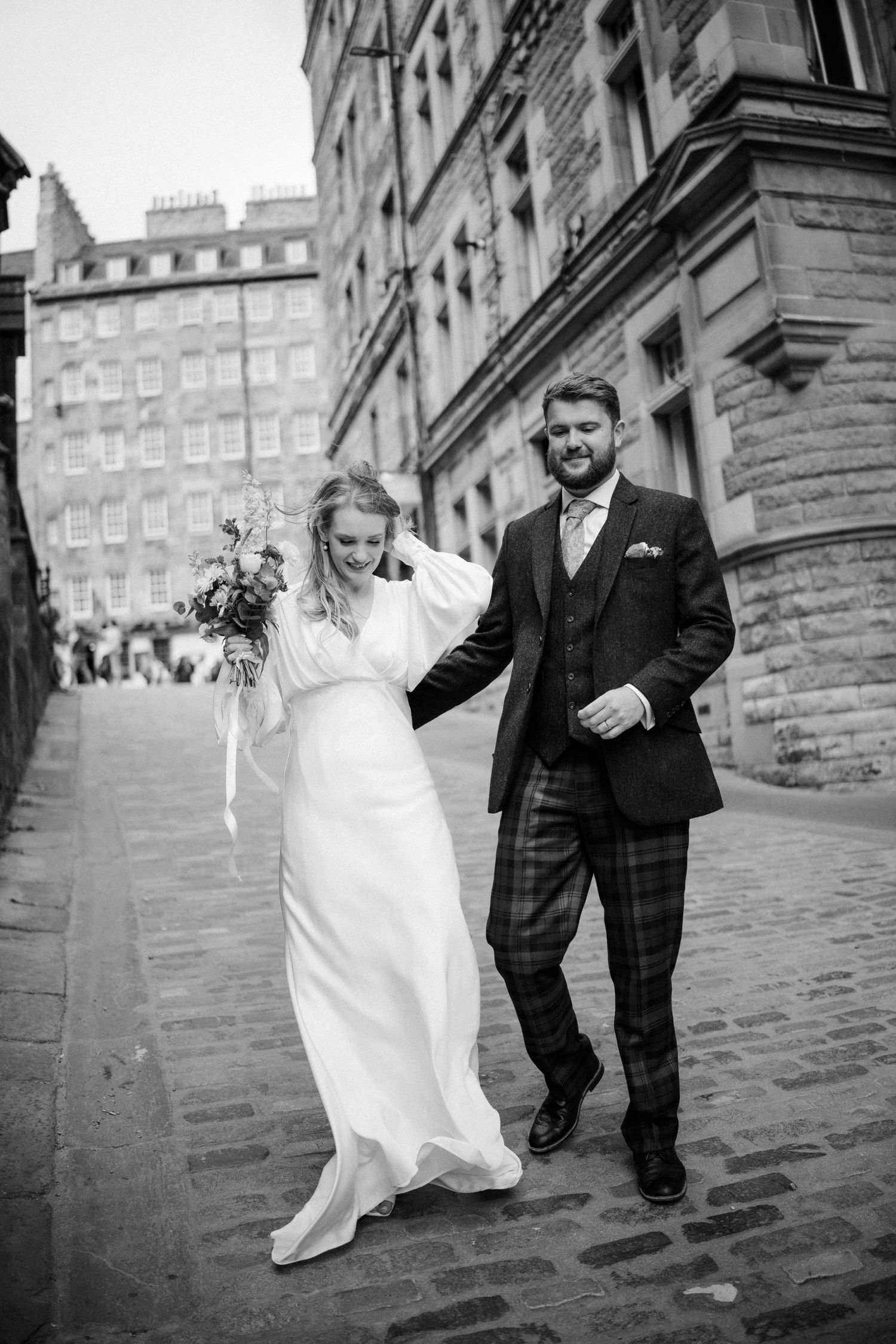 Black and white photo of a smiling bride and groom walking arm in arm on a cobblestone street.