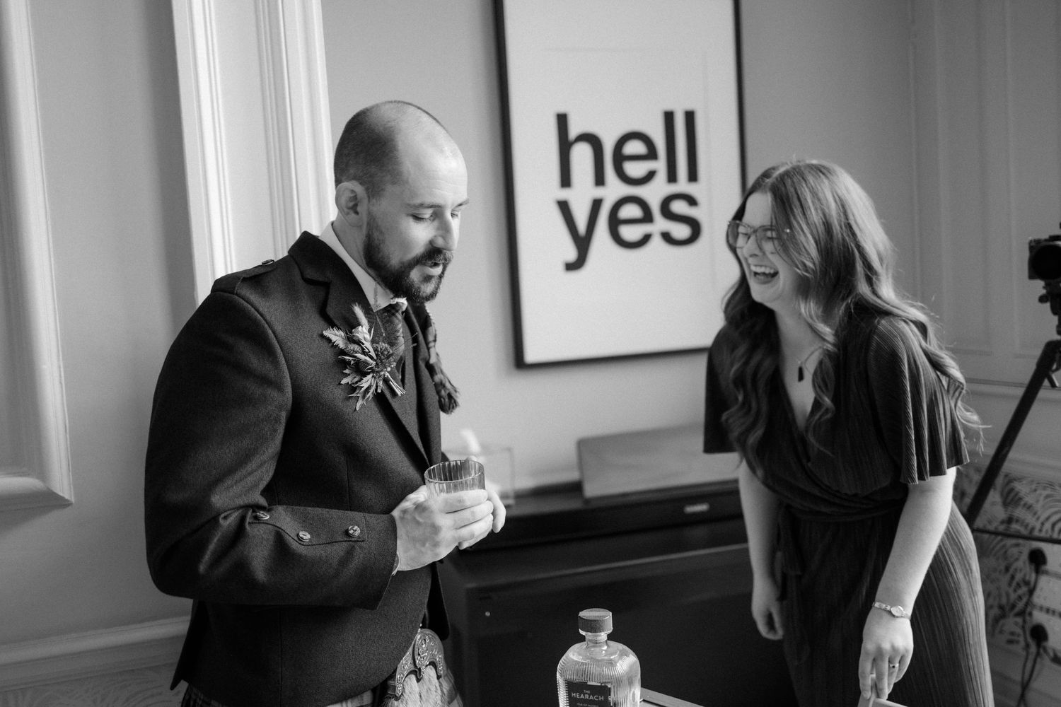 Black and white photo of groom in kilt laughing with woman near a bold hell yes wall sign.