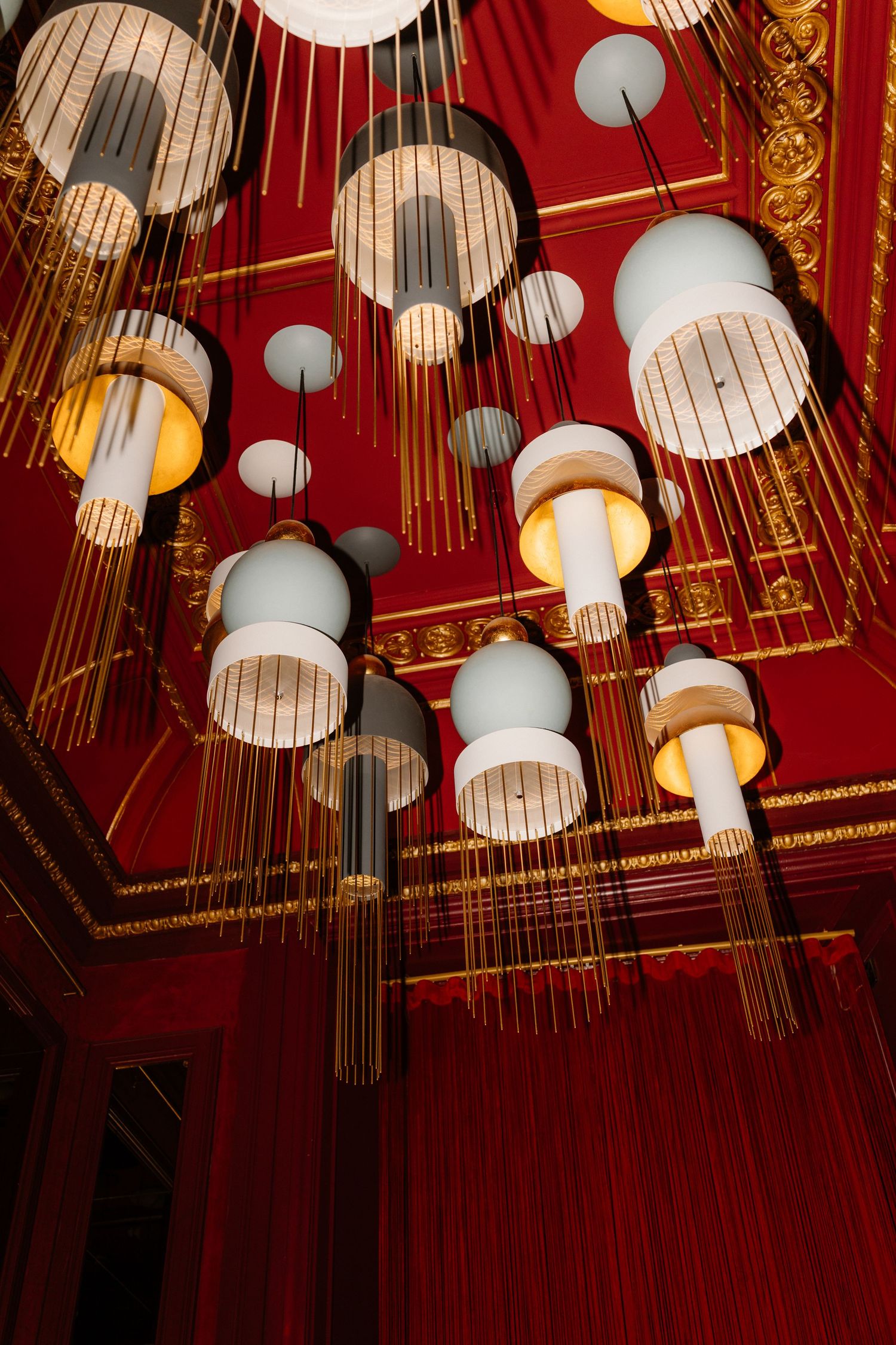 Elegant chandelier installation with cylindrical pendant lights hanging from ornate red and gold ceiling.