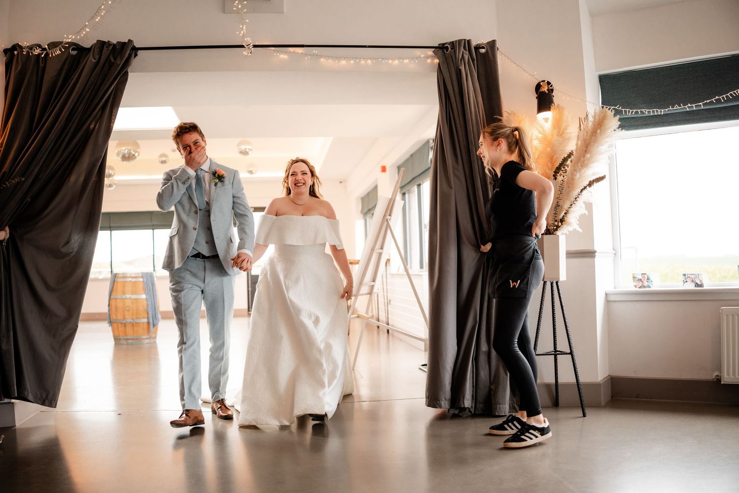 Bride and groom make grand entrance at wedding venue, holding hands, as assistant holds back dark curtain drape.